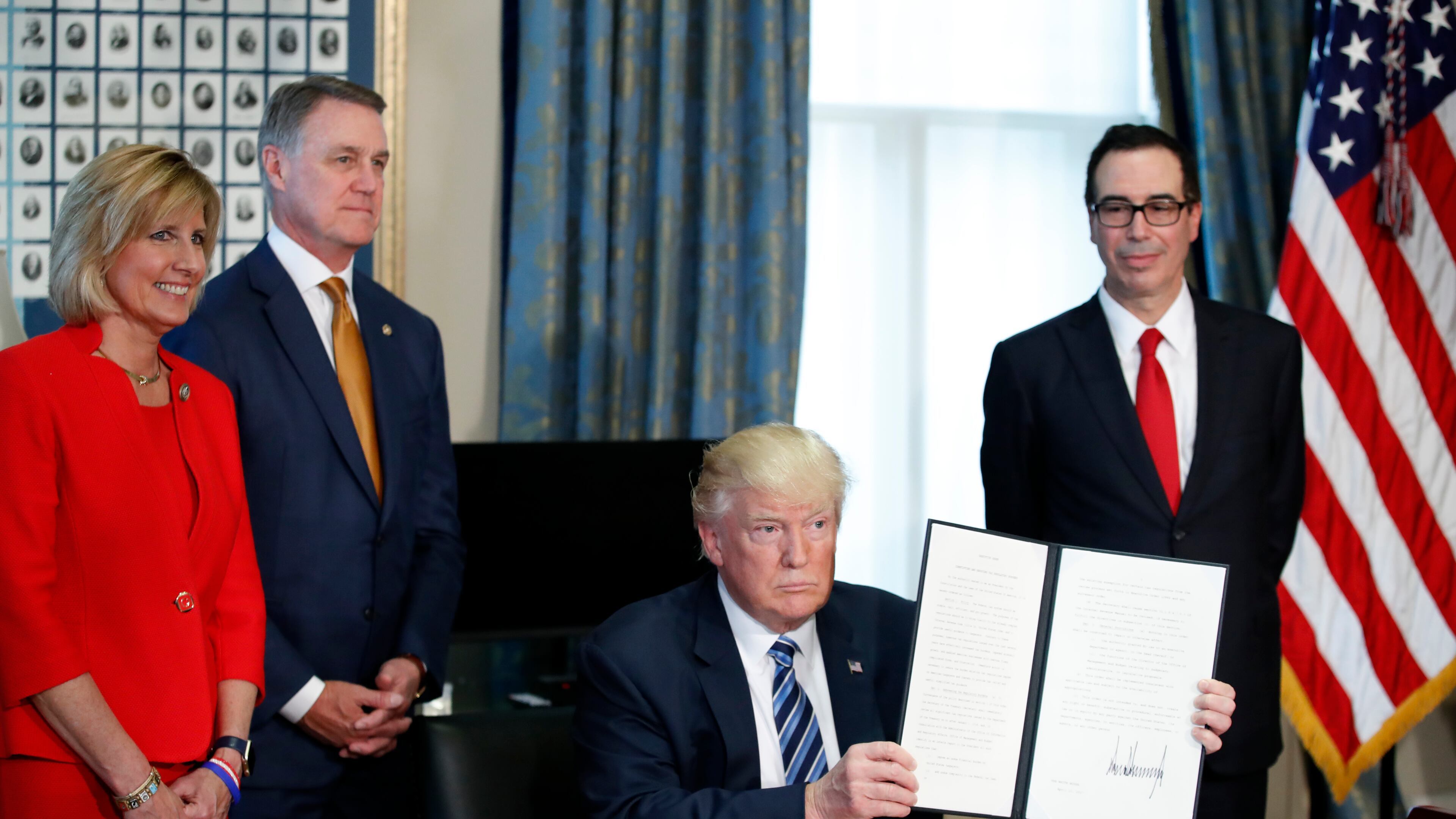 President Donald Trump, accompanied by, from left, Rep. Claudia Tenney, R-N.Y., Sen. David Perdue, R-Ga., and Treasury Secretary Steve Mnuchin, holds up a signed Executive Order, Friday, April 21, 2017, at the Treasury Department in Washington. (AP Photo/Alex Brandon)