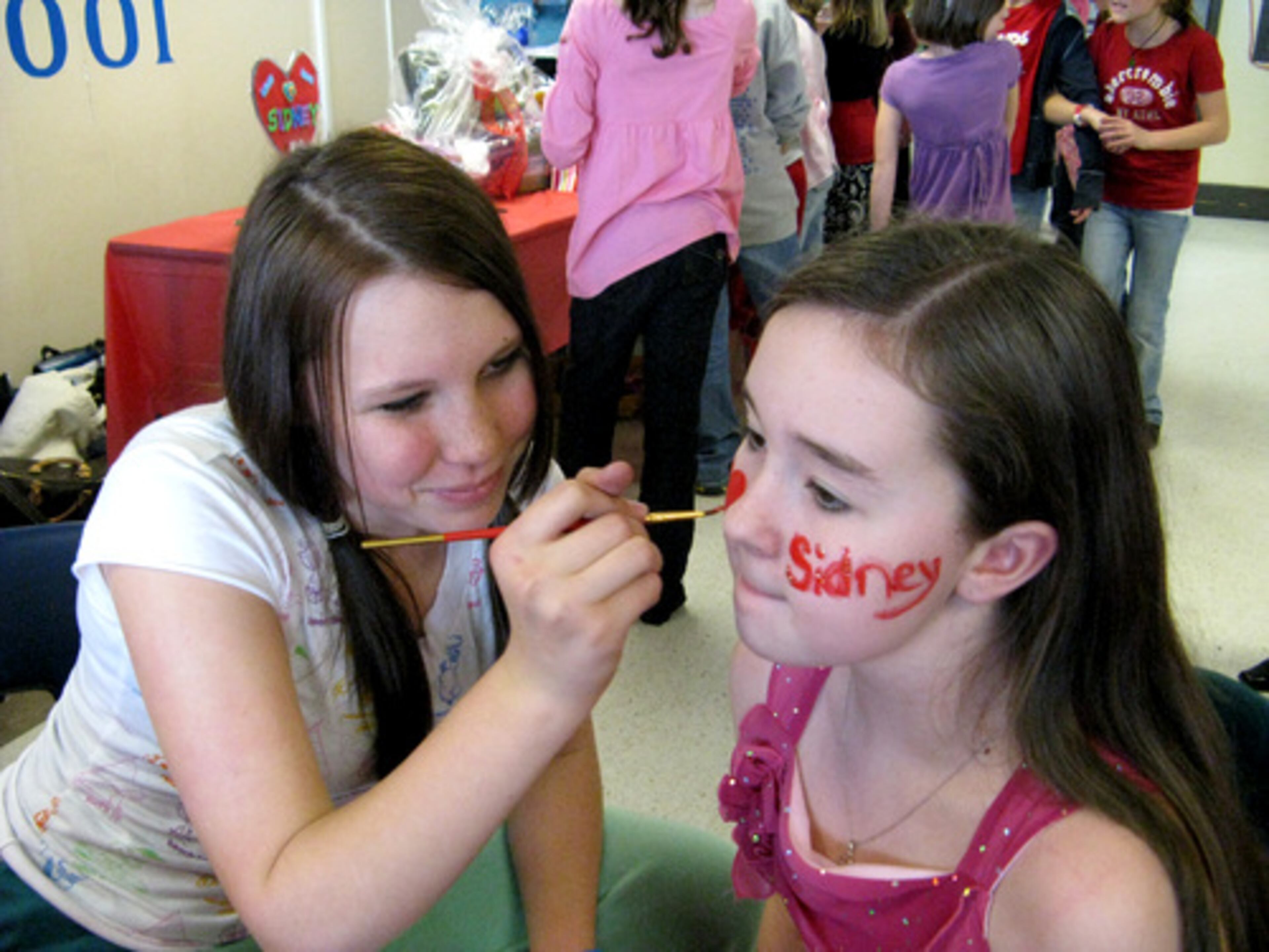 Eighth-grader Olivia Lathem paints "I (heart) Sidney" on Sara Owens' face.
