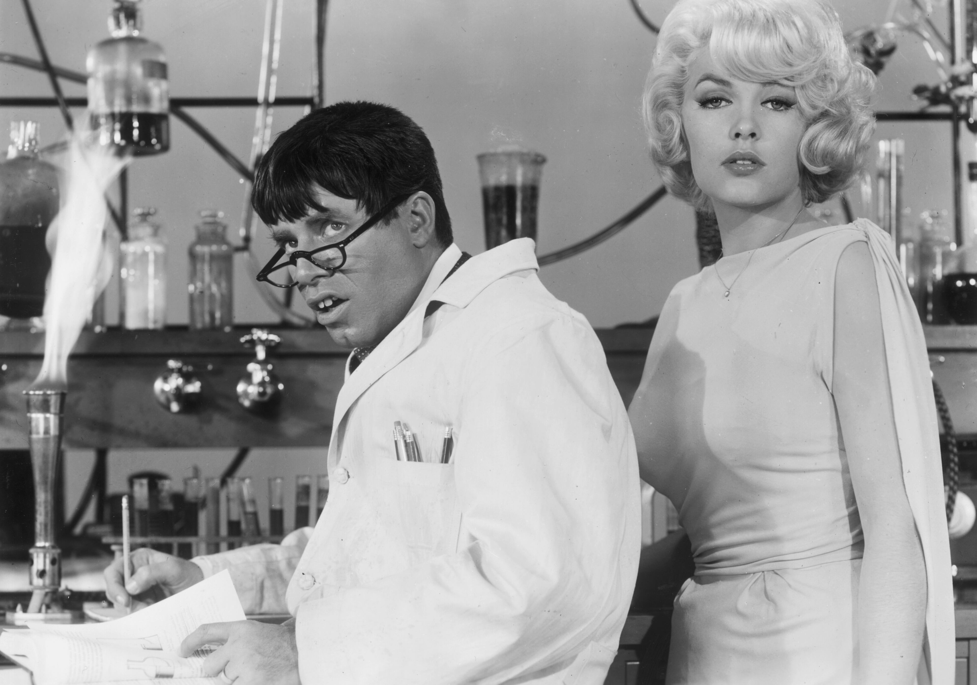 1963: American actor and comedian Jerry Lewis wearing a lab coat and glasses, as actor Stella Stevens poses next to him, in a still from Lewis's film, 'The Nutty Professor'. (Photo by Paramount Pictures/Getty Images)