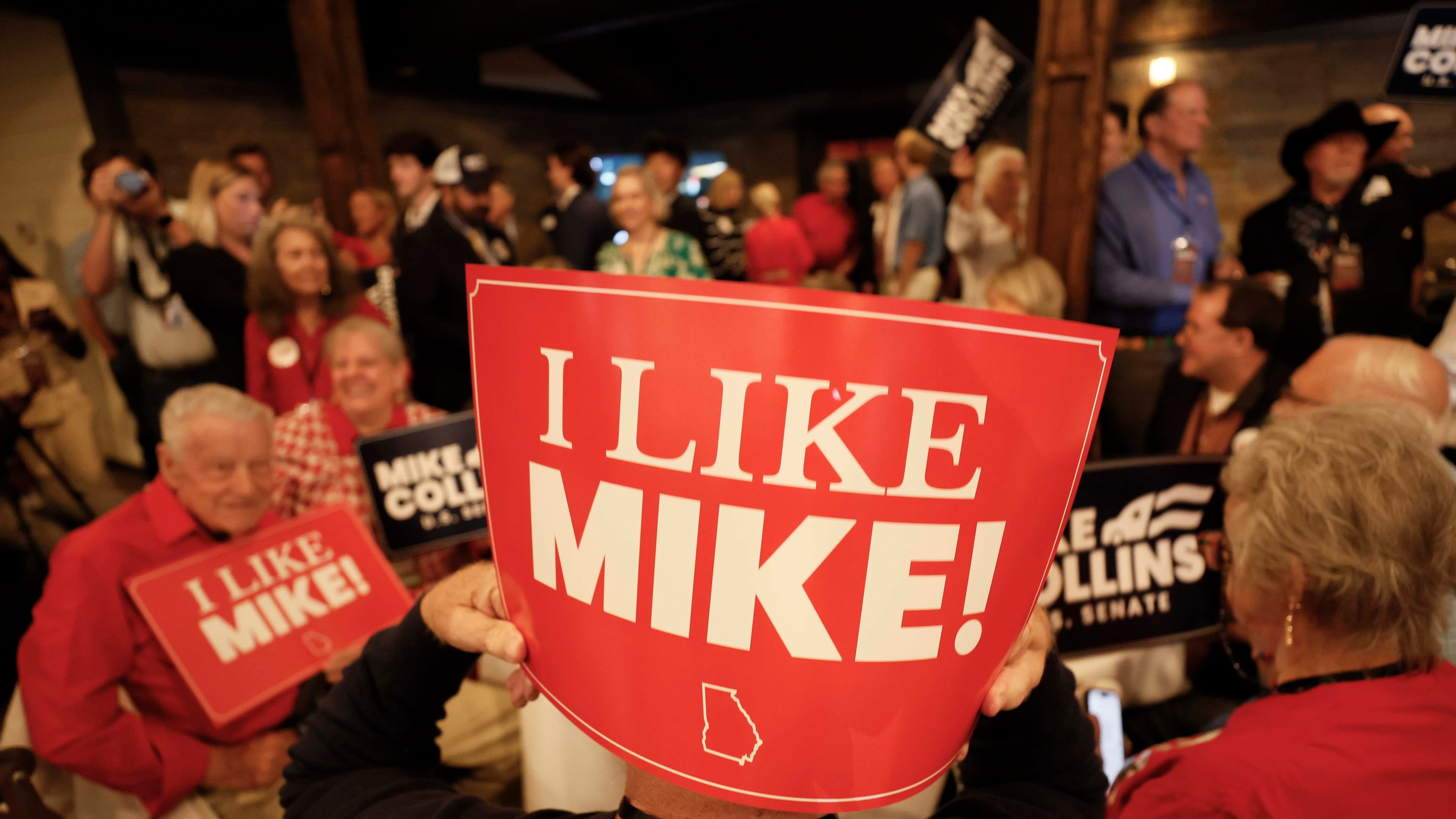 Supporters of U.S. Rep. Mike Collins hold banners as they watch him arrive on stage for his speech during the runoff election watch party in Jackson last month. (Miguel Martinez/AJC)