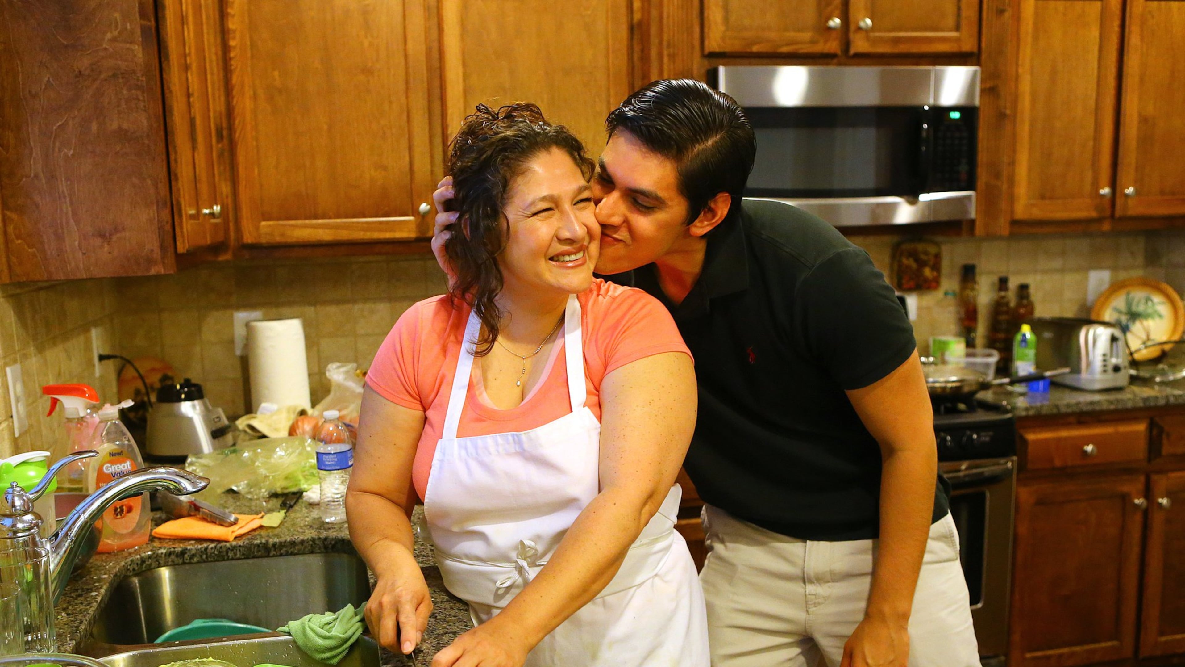 Christian Jimenez gives his mother Maria Velasquez a kiss on the cheek while helping her prepare the family dinner in the kitchen of the family home recently in Buford. CURTIS COMPTON / CCOMPTON@AJC.COM