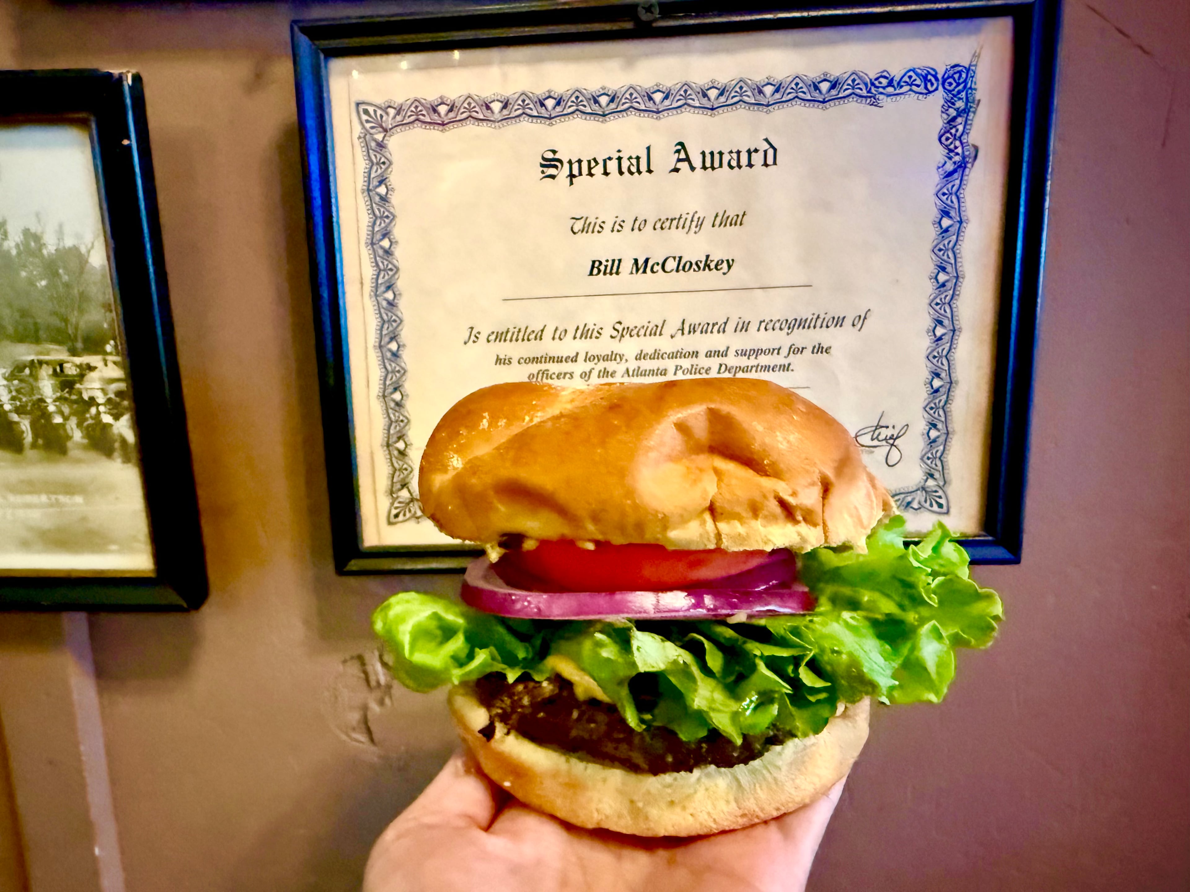 Journalist Angela Hansberger holds the McCloskey cheeseburger in front of the sign honoring the longtime Manuel's Tavern bartender Bill McCloskey. (Angela Hansberger for the AJC)