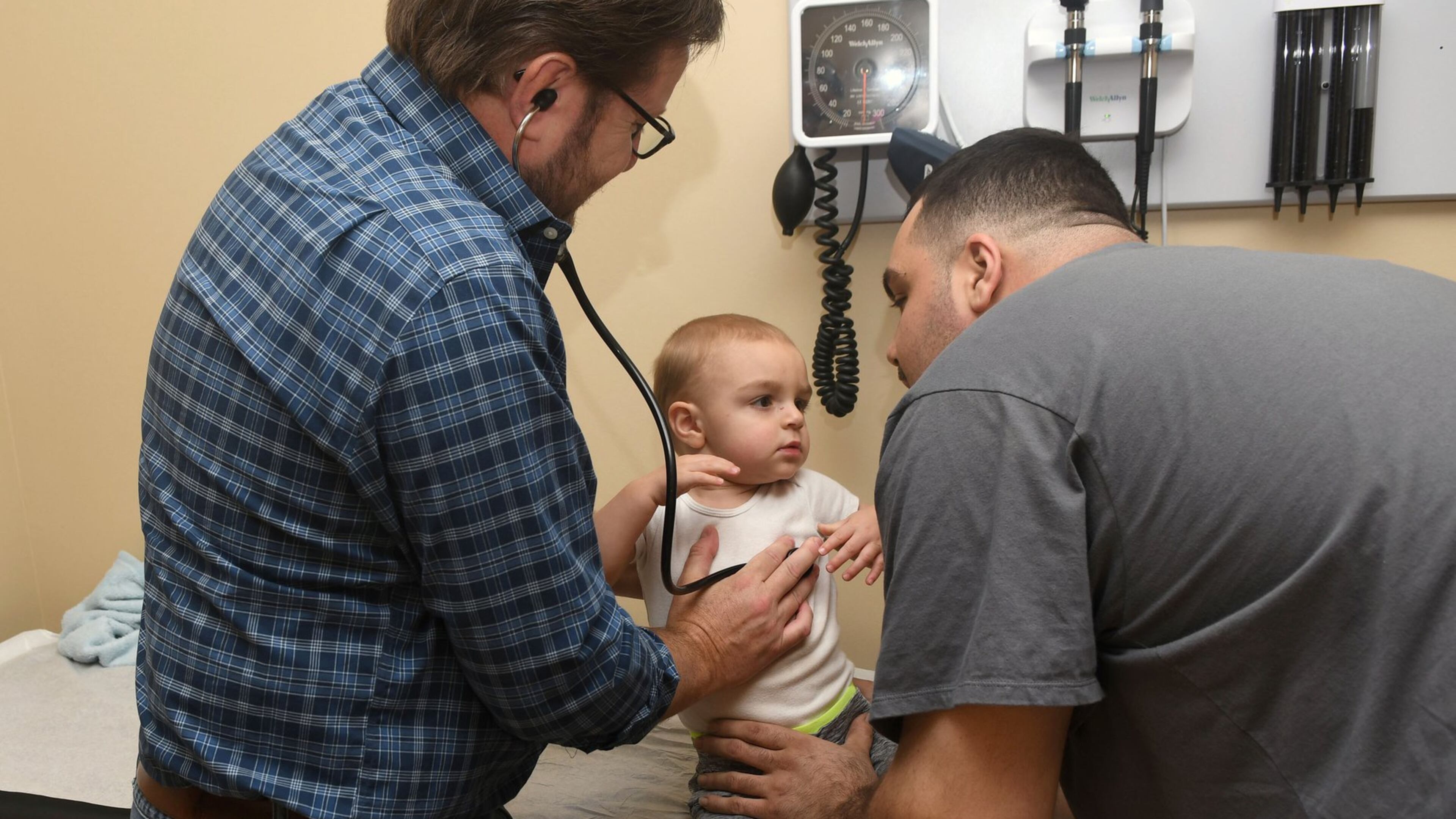 Dr. Greg McHan, in the checked shirt and jeans, examines Lenny Falletta III, 18 months, of Blairsville with a little help from Lenny Falletta Jr., his dad. They are at the Georgia Mountains Health clinic in Blue Ridge. Georgia Mountains Health needs to add another pediatrician, which is not easy in a rural area. But as Congress let the national clinic program’s funding expire, Georgia Mountains has put the brakes on attending recruiting fairs and hiring a recruitment firm. CEO Steve Miracle said he “can’t in good conscience” say federal funding will be there to sign a multiyear contract. (Photo by Rebecca Breyer / special to the AJC)