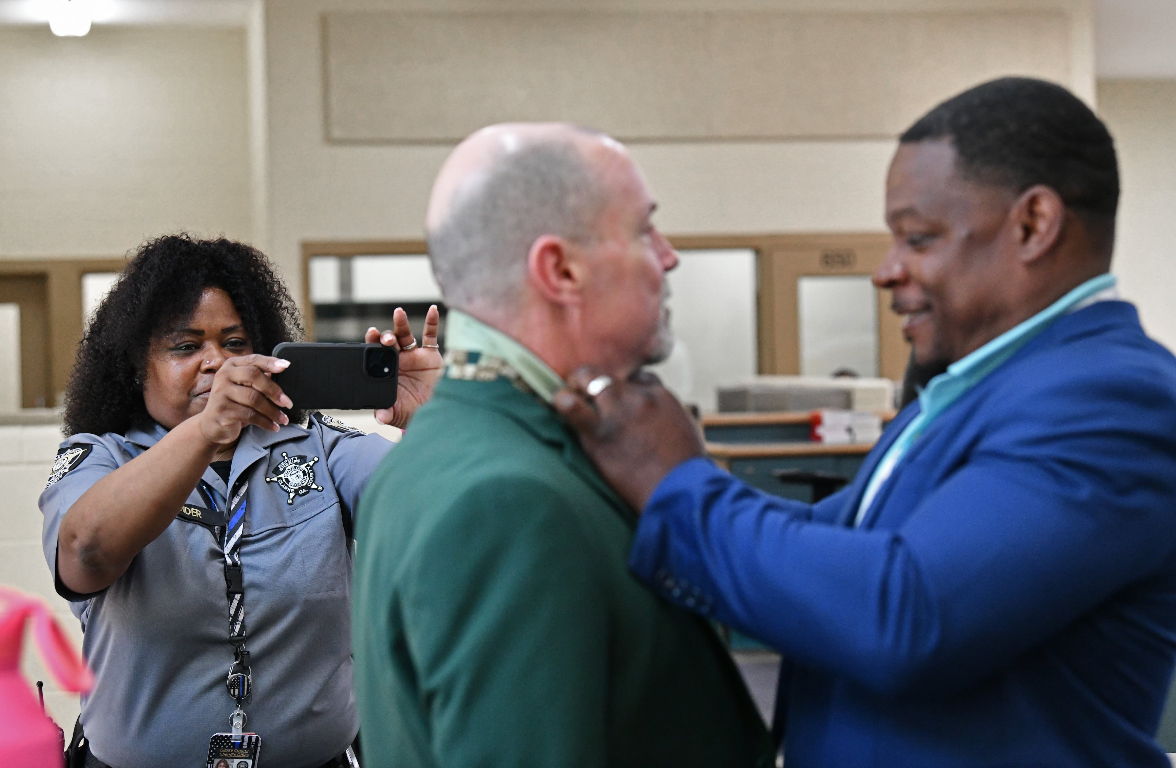 Deputy Gloria Pender (background) takes a picture as Shane Sims (right), founder of Principles over Passion, helps graduate Kirby Kesler with a necktie before their graduation ceremony for the Re-Entry Success Program at the Athens-Clarke County Jail on Wednesday, June 18, 2025, in Athens. (Hyosub Shin/AJC)