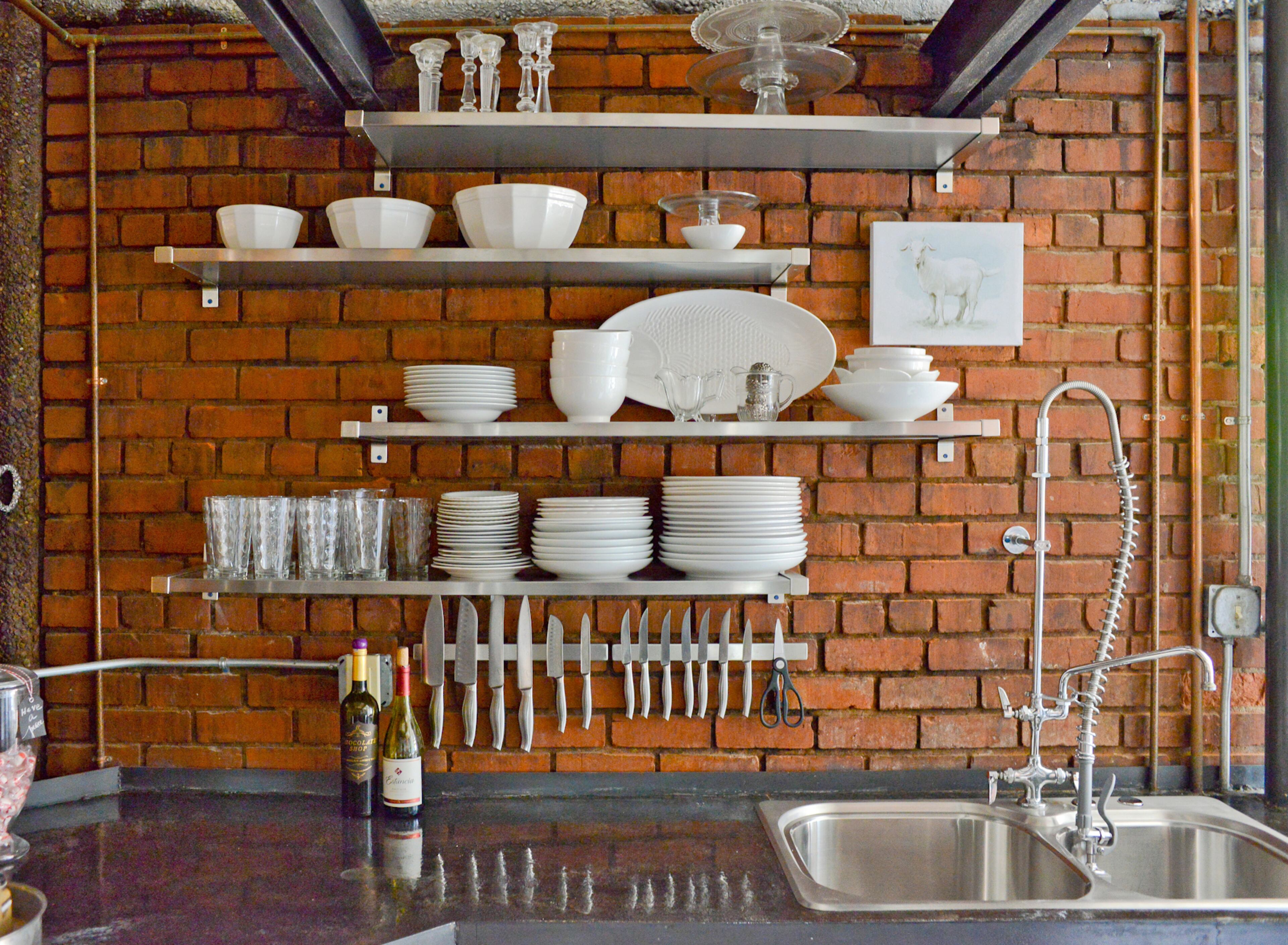 Open metal shelving puts white dinnerware, knives and glasses within easy reach and as a contrast to the exposed brick walls. At the top, you can see the original trolley system used when the building was a meatpacking factory.