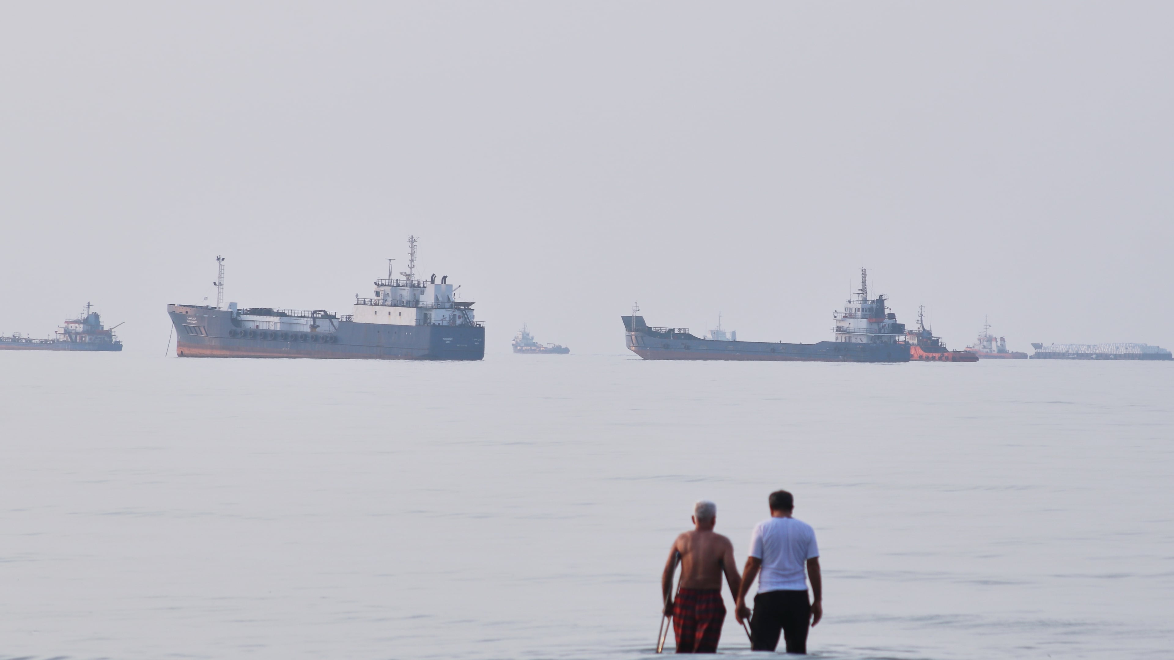 Two men wade in the waters of the Strait of Hormuz with vessels anchored in the background, off Bandar Abbas, Iran, Sunday, July 12, 2026. (Razieh Poudat/ISNA via AP)