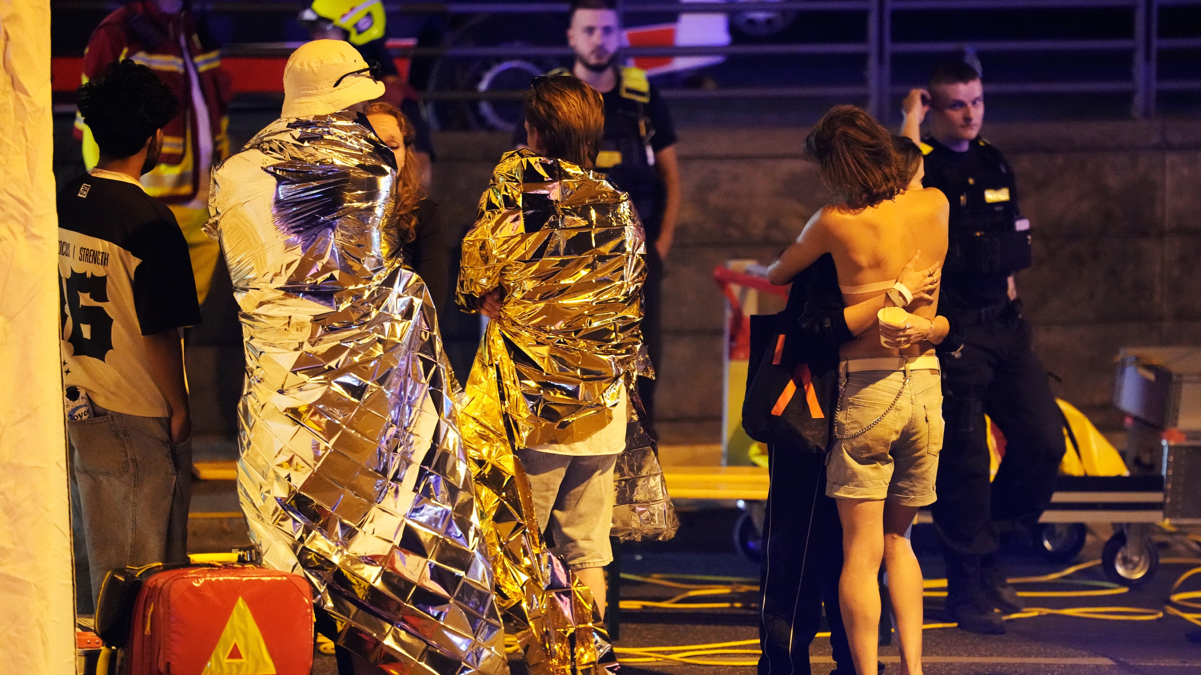 Two people embrace near others wearing thermal blankets as they stand near emergency personnel in Berlin, early Sunday, July 26, 2026, following the cancellation of Christopher Street Day festivities. (AP Photo/Markus Schreiber)