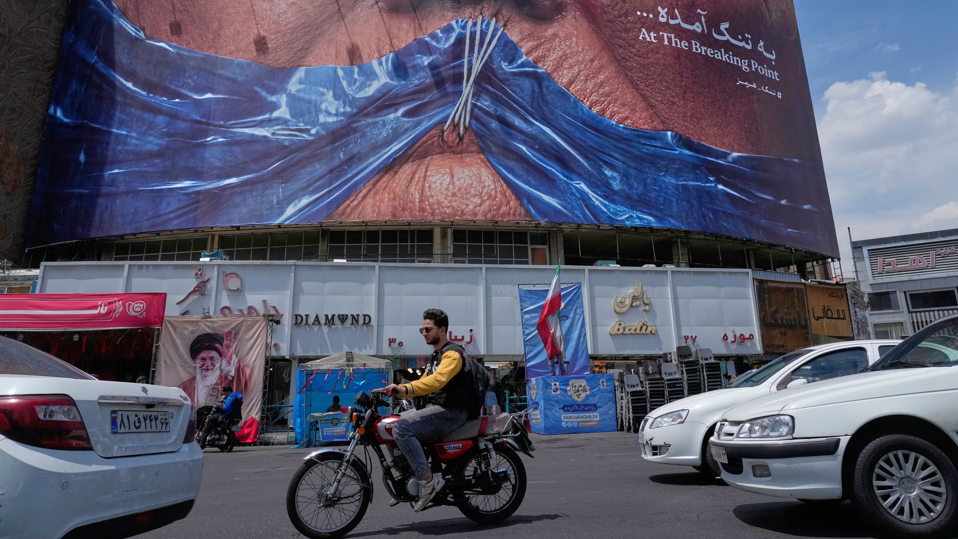 Vehicles drive past a billboard with graphic showing Strait of Hormuz and sewn lips of U.S. President Donald Trump in a square in downtown Tehran, Iran, Saturday, May 2, 2026. (AP Photo/Vahid Salemi)