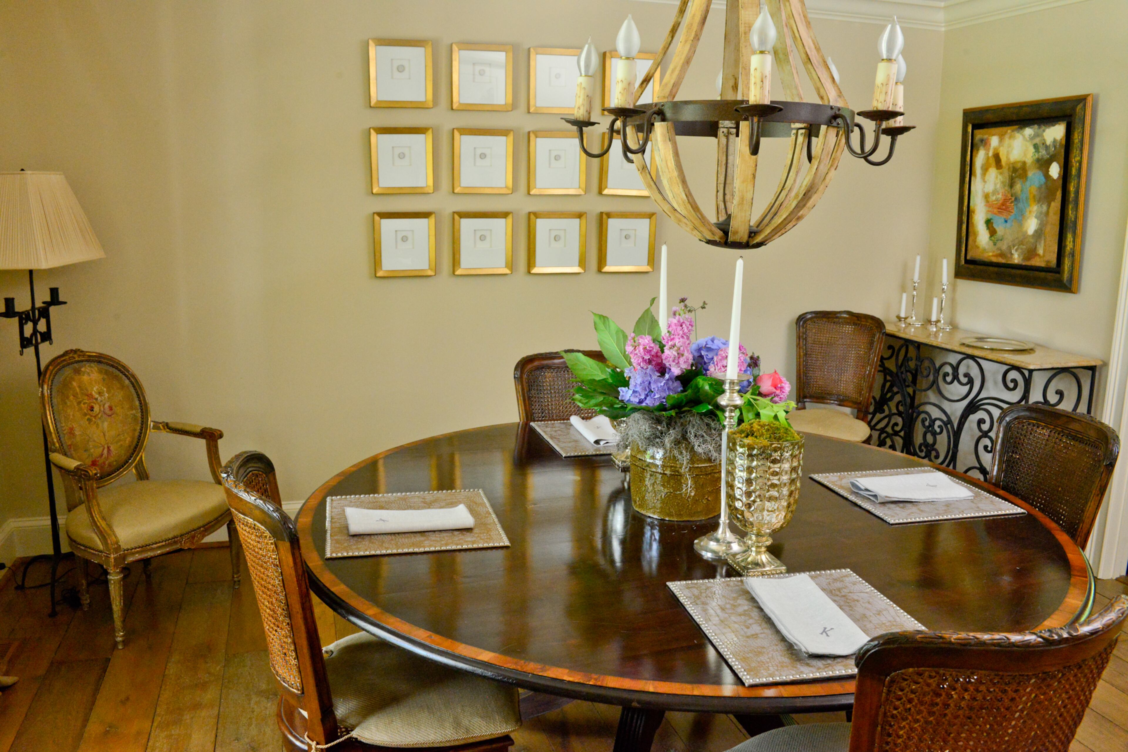 A light wood chandelier from Boxwoods accents the darker wood of the round table in the home's formal dining room. Framed artwork and flowers add splashes of color to the earth-toned room. The artistic wrought iron and wood-topped table is from Boxwoods as well.