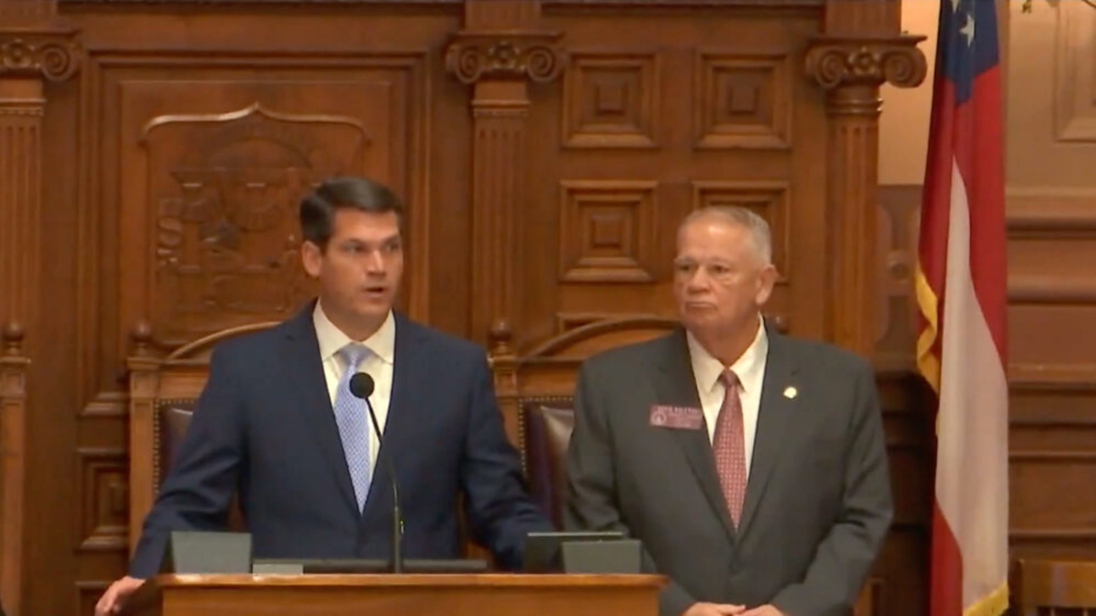 (left) white man standing at podium with navy jacket and blue tie. (right) white man with short white hair with grey jacket, red tie.