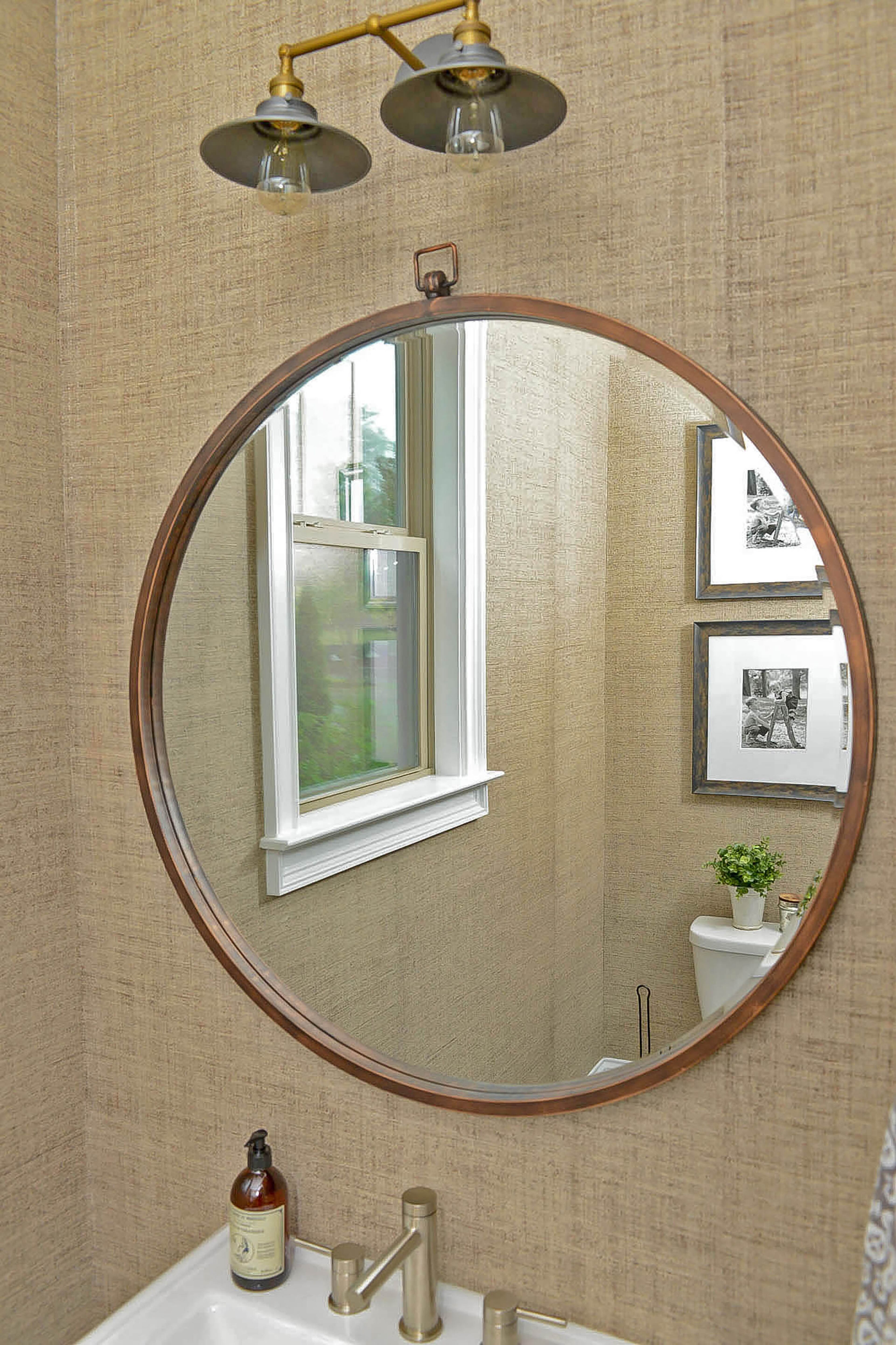 The framed photographs on textured wallpaper, a double vintage and industrial-style sconce from RH with filament bulbs, and a round, metal mirror show off the home's transitional urban style in one of the bathrooms.