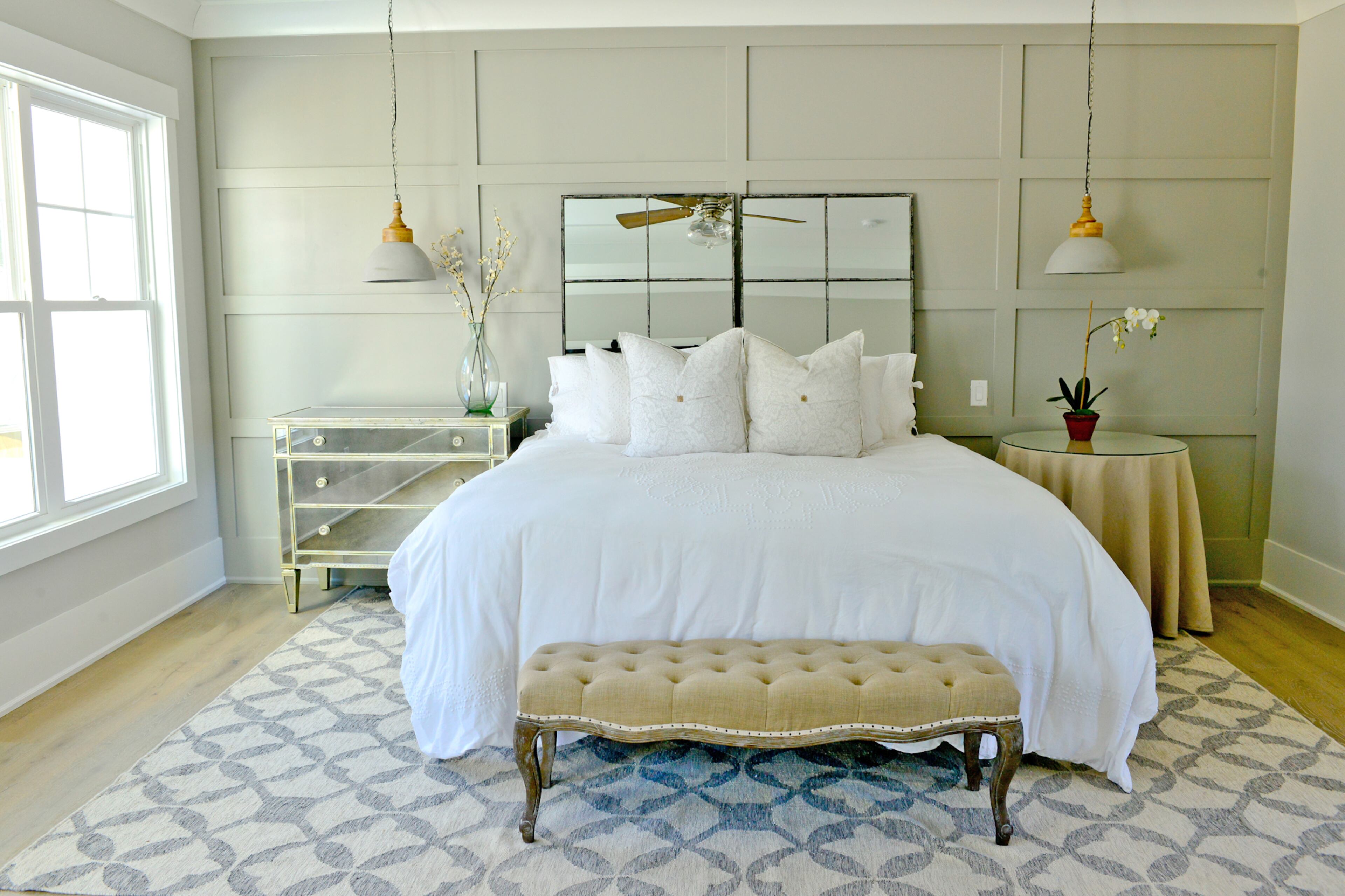A paneled accent wall, painted in Sherwin-Williams Dorian Gray, and mirrored accents like the Horchow bedside dresser and mirrored headboard, add depth to the master bedroom. A gray and white rug from West Elm remains true to the color scheme and anchors the room, while cement pendant lighting from Four Hands creates a sense of symmetry.