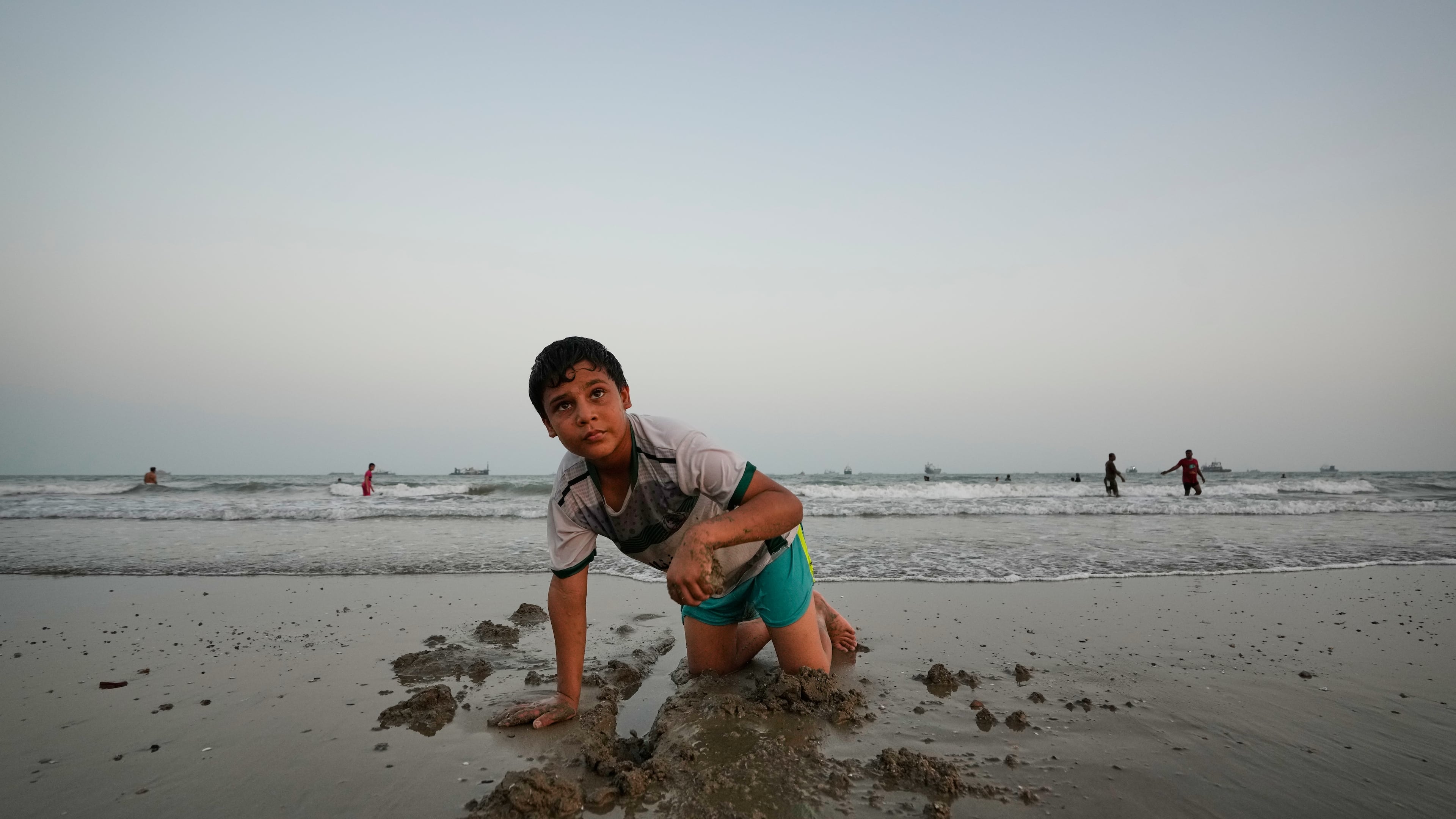 A boy plays in wet sand as others swim and commercial vessels lie anchored in the Strait of Hormuz off Bandar Abbas, Iran, Friday, Sept. 4, 2026. (AP Photo/Vahid Salemi)