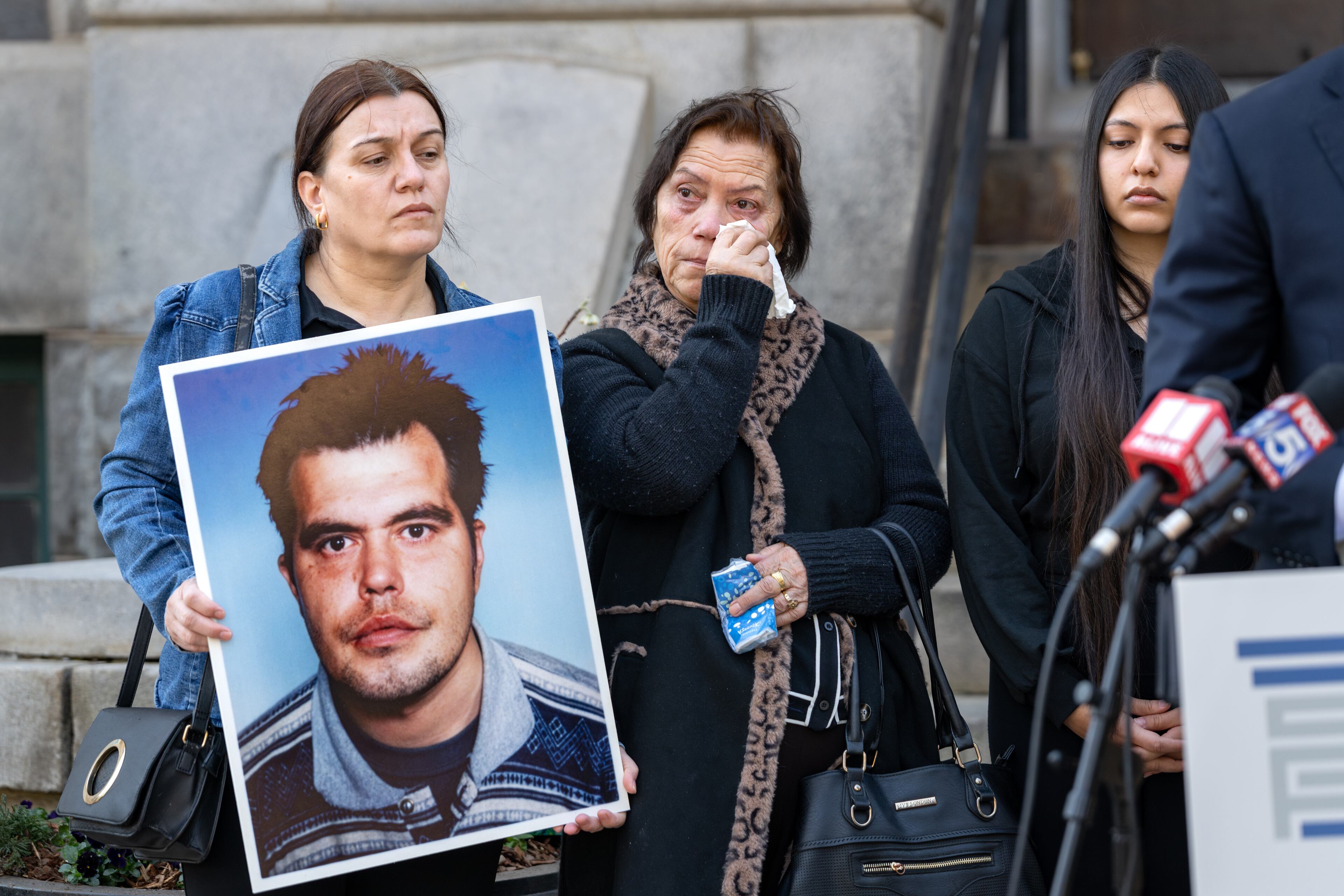 Arben Pilici's mother, Sebahate Pilici (center), wipes away tears at a press conference on Friday, Feb. 13, 2026 to announce a lawsuit against the DeKalb County Police Department in the death of her son. Arben's sister Drandofile Pilici holds a photo of him. They are joined by Arben's niece Lorita Jashari. (Ben Hendren for the AJC)