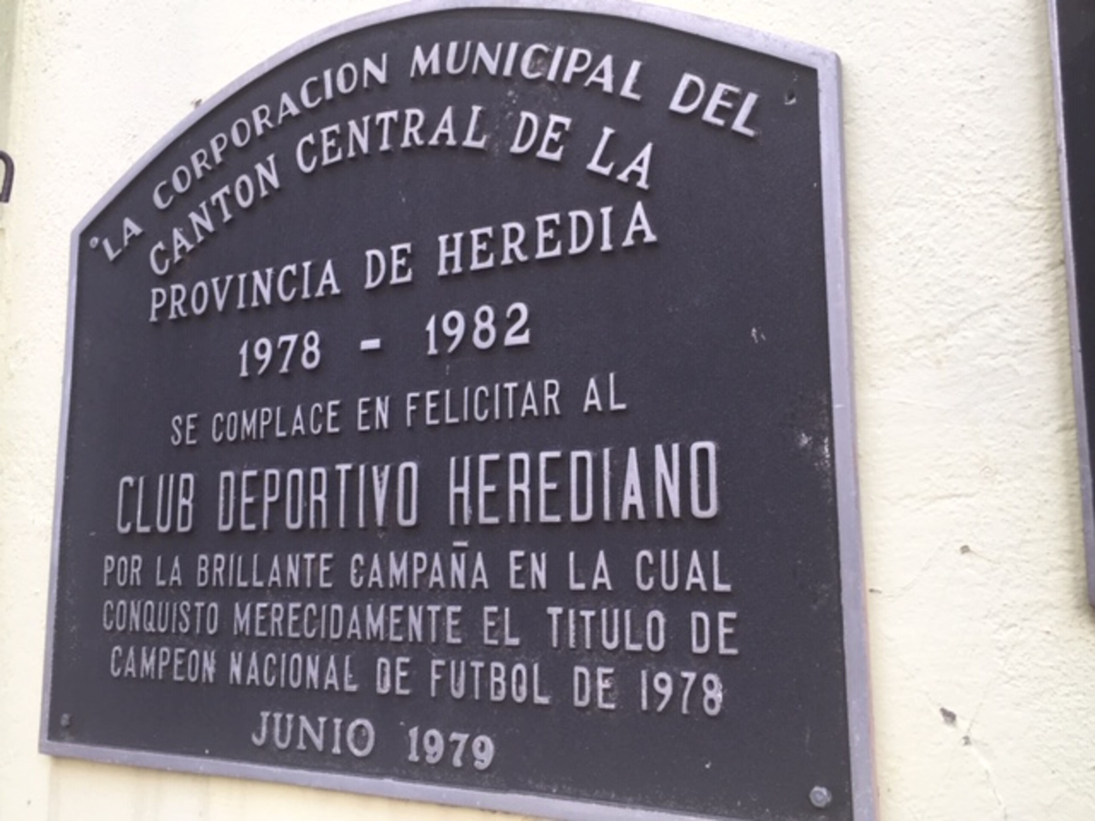 A plaque commemorating the stadium and team is affixed to a wall that you walk by as you enter the stadium from a corner near the street. The field is ahead.