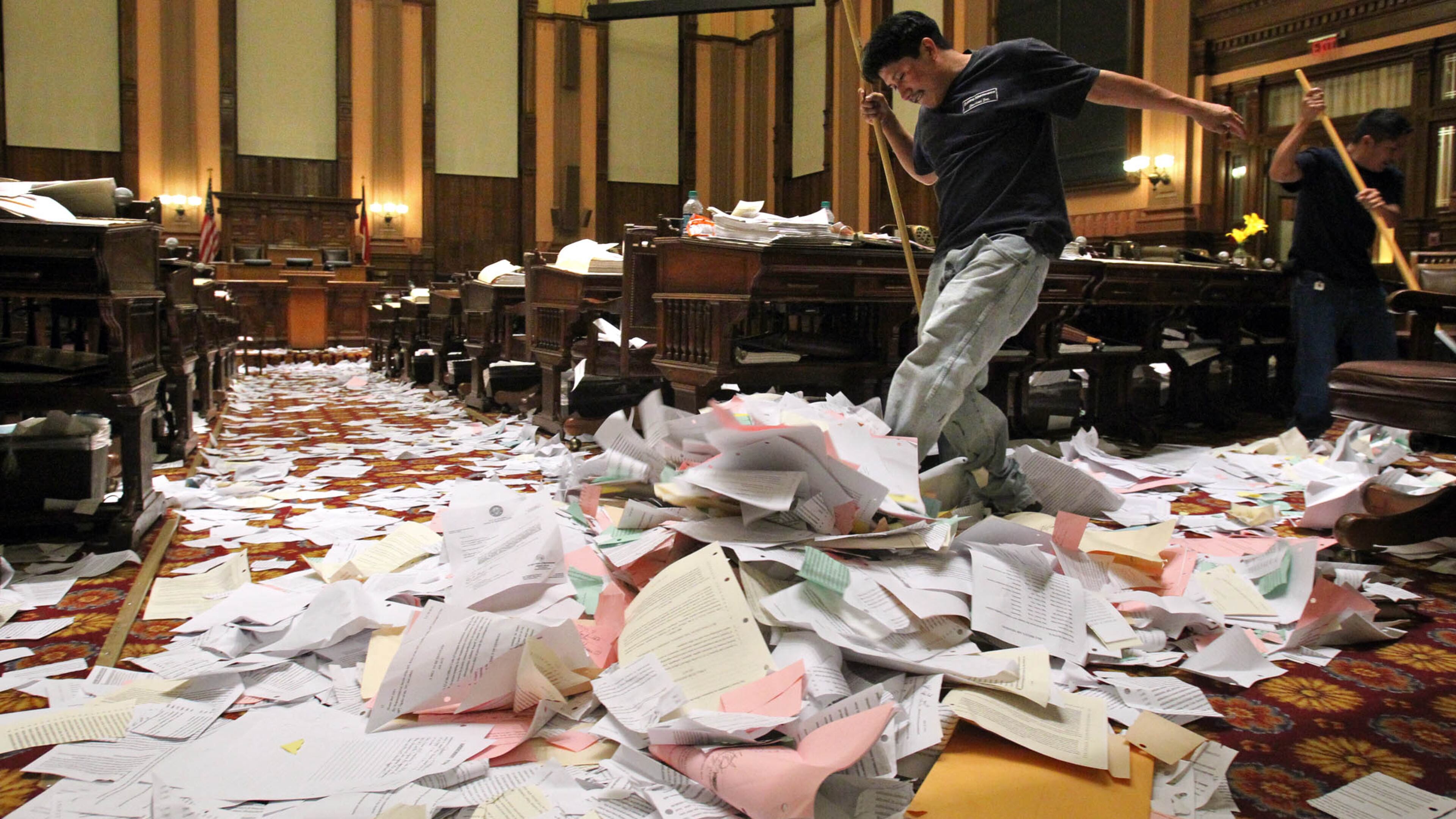 Building Maintenance Service employee Nelson Alisea kicks a pile of paper toward the center aisle as he and others start the clean up after, "Sine Die," in the House of Representatives after the last Legislative Day of the 2012 session at the Capitol early Friday morning.