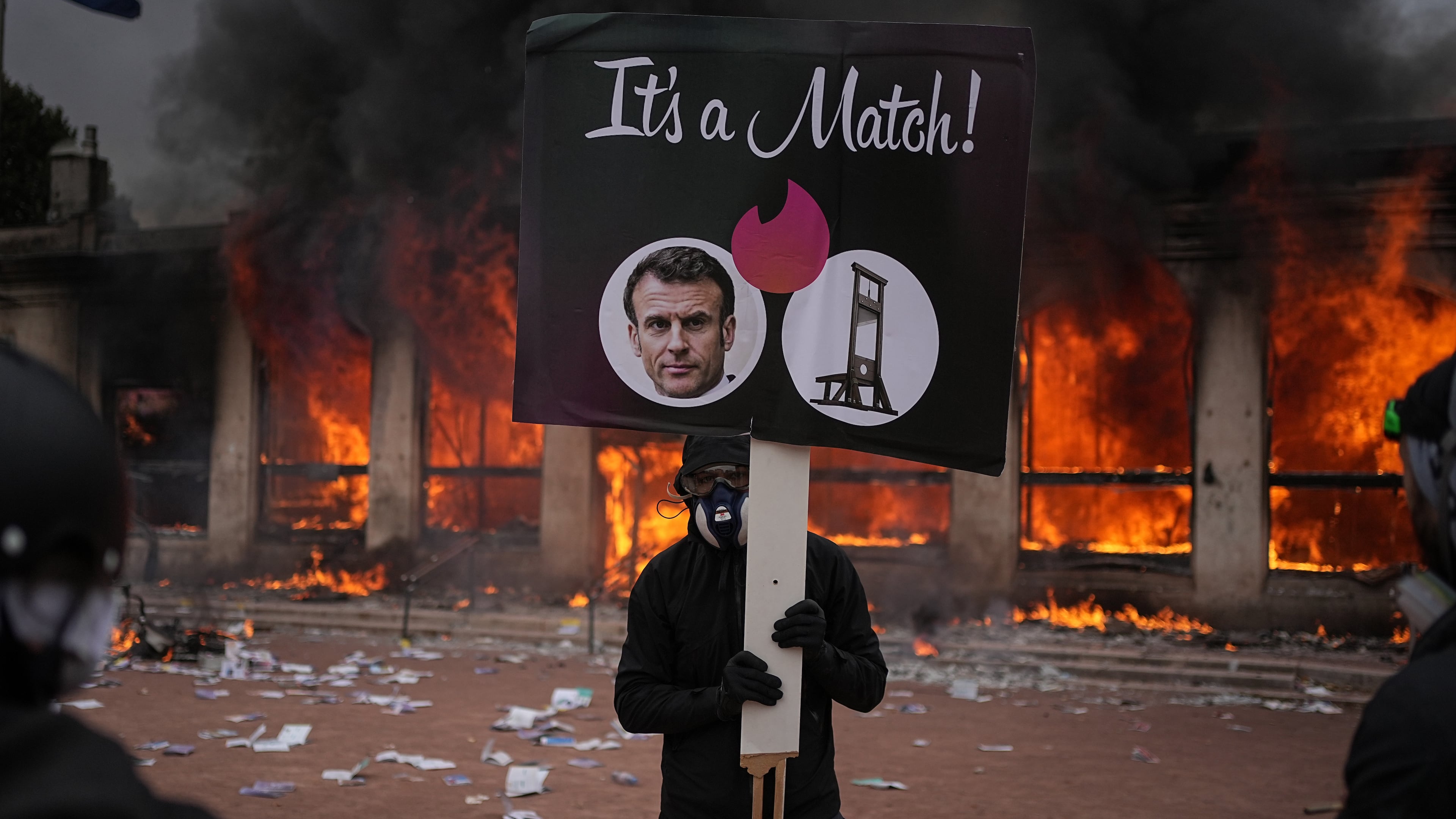 A protester holds a placard with a photo of French president Emmanuel Macron and a guillotine in front of a burning tourism office during a demonstration against rising living costs and in solidarity with students' grievances in Lyon, France, Saturday, Oct. 17, 2026. (AP Photo/Laurent Cipriani)