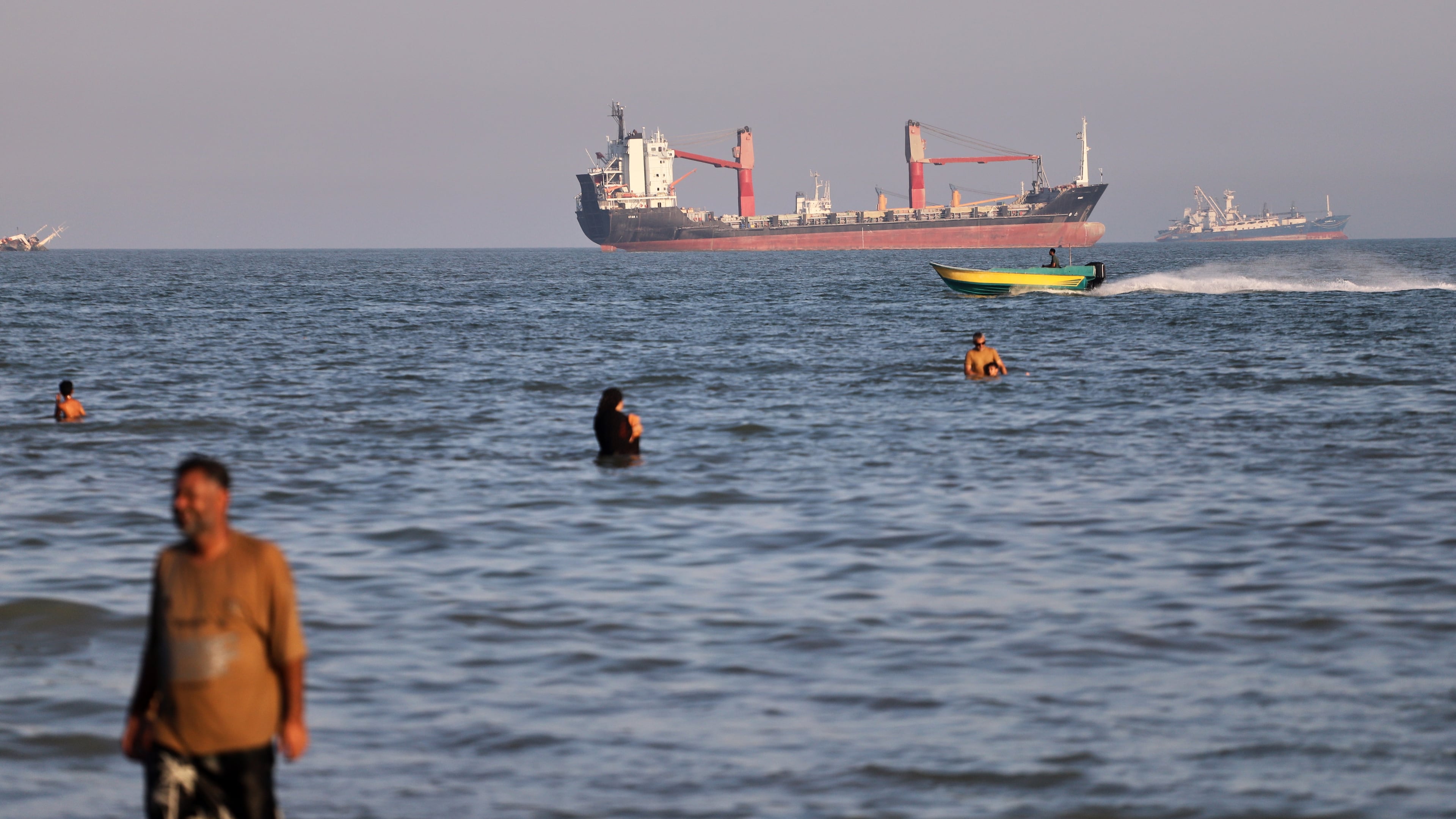 Residents swim in the waters of the Strait of Hormuz as a small motorboat passes cargo ships and other commercial vessels offshore near Bandar Abbas, Iran, Wednesday, June 17, 2026. (Amirhosein Khorgooi/ISNA via AP)