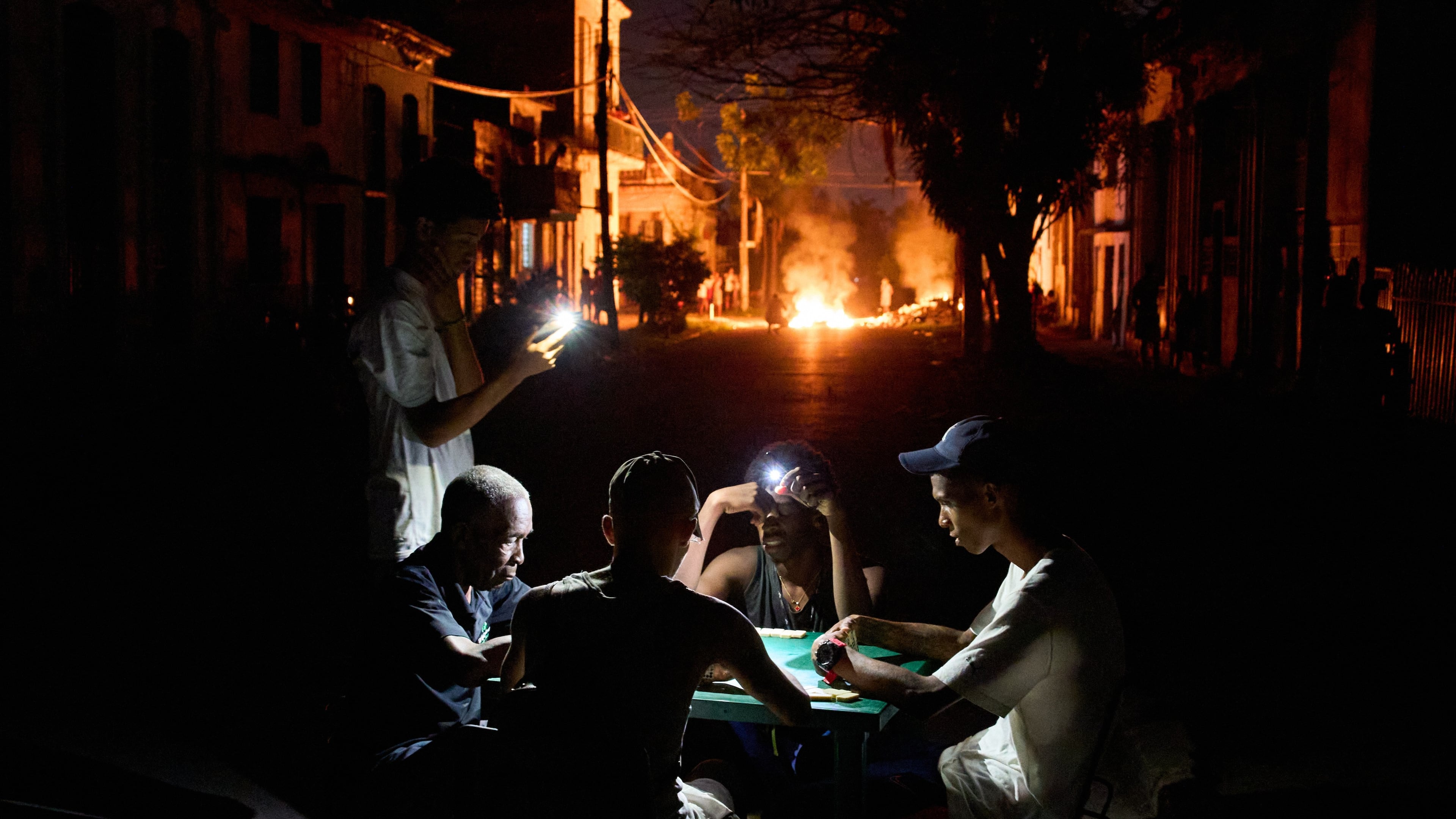 People illuminate themselves with their phones while playing dominoes as a fire set up by residents protesting against prolonged power outages burns on a street in Havana, Cuba, Thursday, May 14, 2026. (Ramon Espinosa/AP)