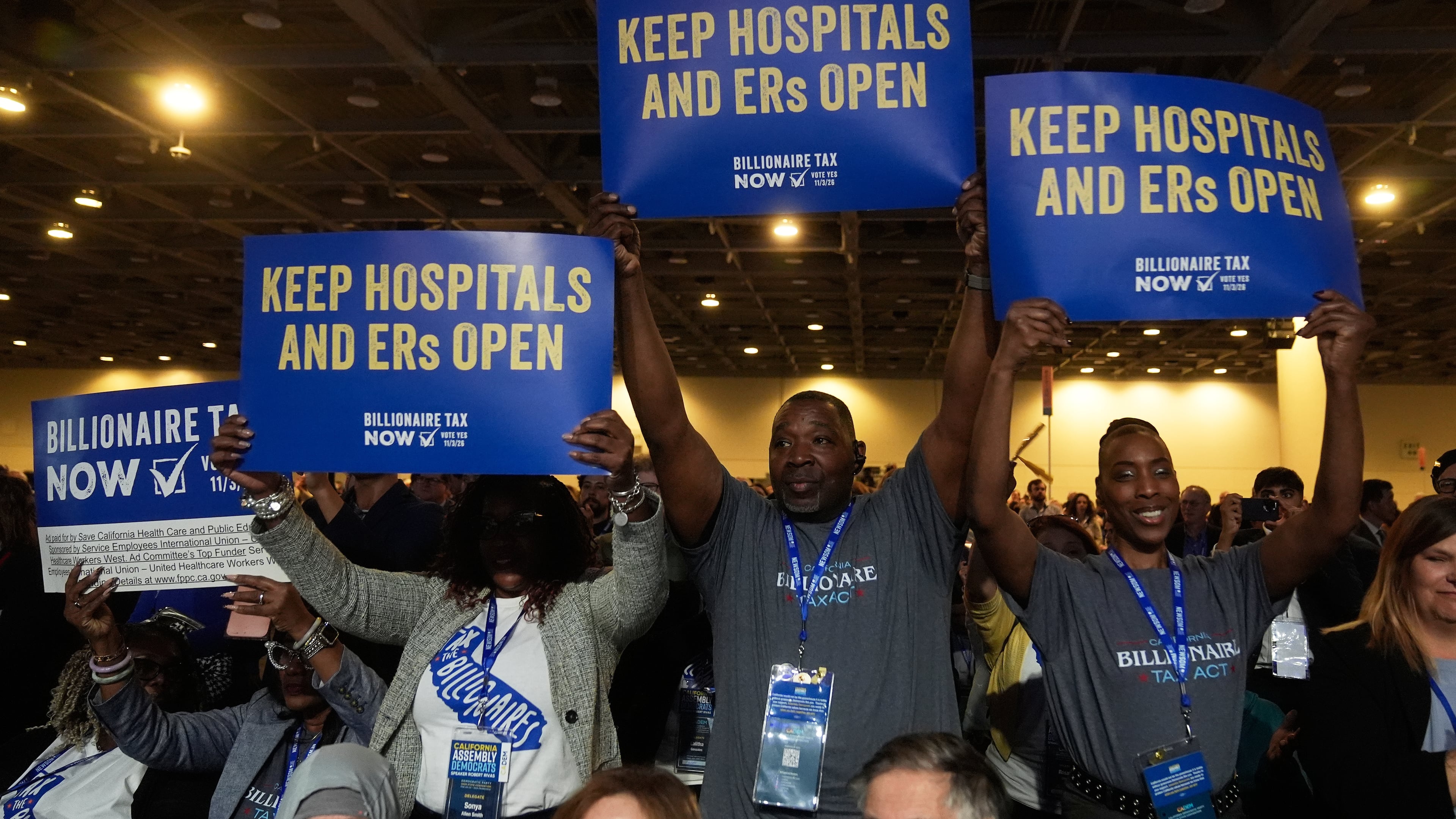 FILE - People supporting Billionaire Tax Now hold up signs at the 2026 California Democratic Party State Convention in San Francisco, Feb. 21, 2026. (AP Photo/Jeff Chiu, File)