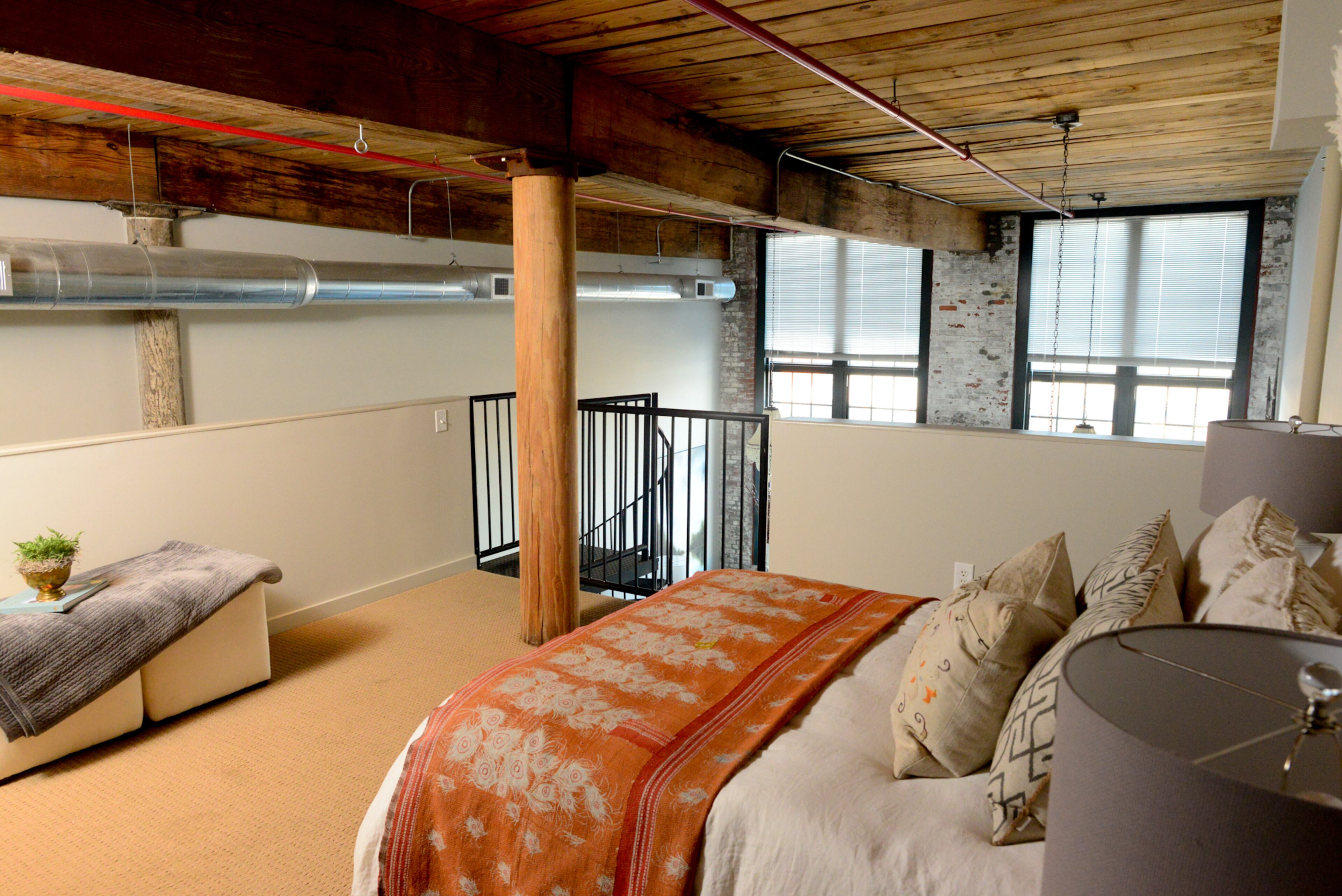 Upstairs, a guest bedroom is surrounded by vintage industrial architectural elements, such as the wood plank ceilings, exposed ductwork and brick walls. The bedding is from Matteo.