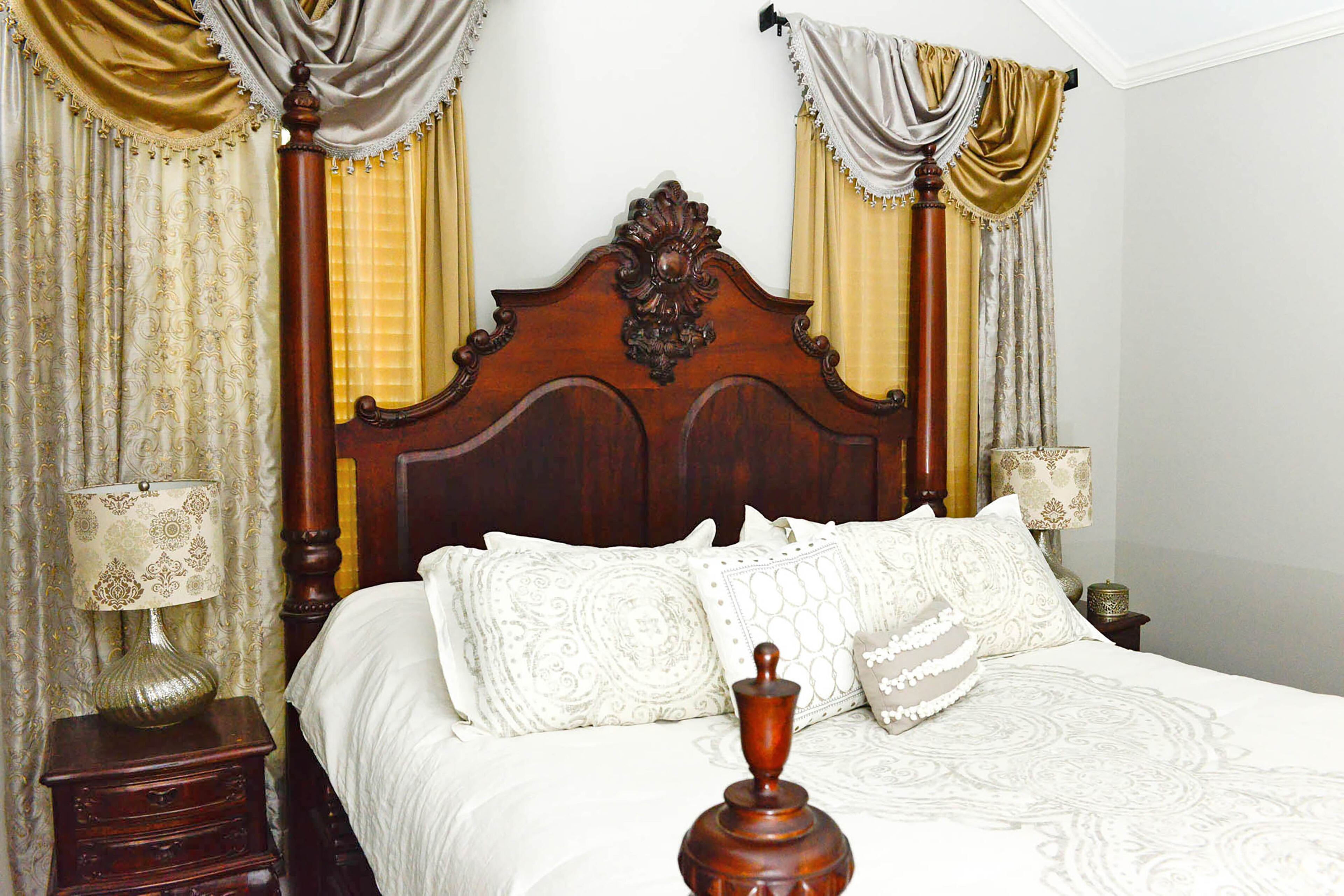 The silver and gold curtains bring out the richness of the wood bed in master bedroom. Walls painted Colonnade Gray, a Sherwin-Williams hue, are a neutral backdrop to the draperies and gold lamps with patterned shades.