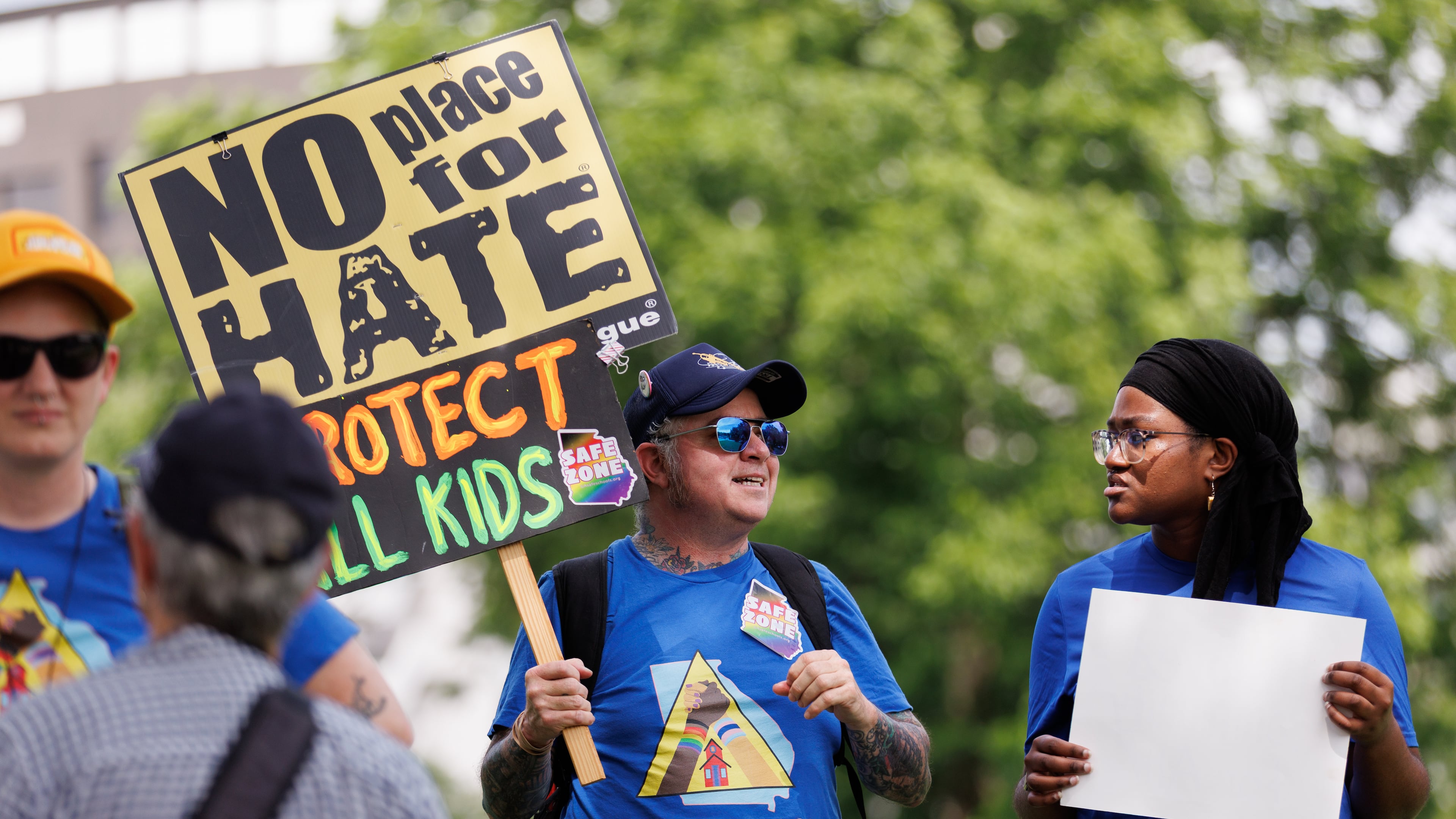A small group of protestors rally before a school board meeting in Decatur on Thursday. SJ, his activist name, wears stickers and a T-shirt showing him as a “safe zone.” Protesters are upset with what they believe is a lack of transparency around building an early learning center and the district's connection to the superintendent's controversial podcaster husband. (Estela Muñoz/AJC)