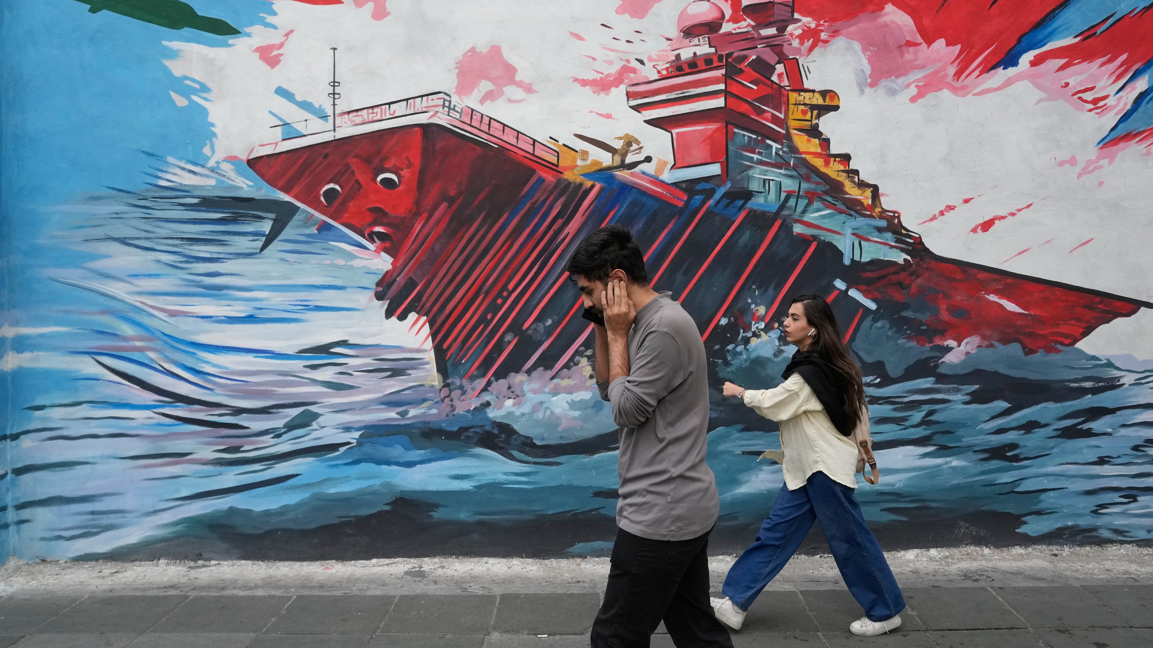 People walk past a mural depicting a U.S. aircraft carrier under missile attack in downtown Tehran, Iran, Sunday, May 17, 2026. (AP Photo/Vahid Salemi)