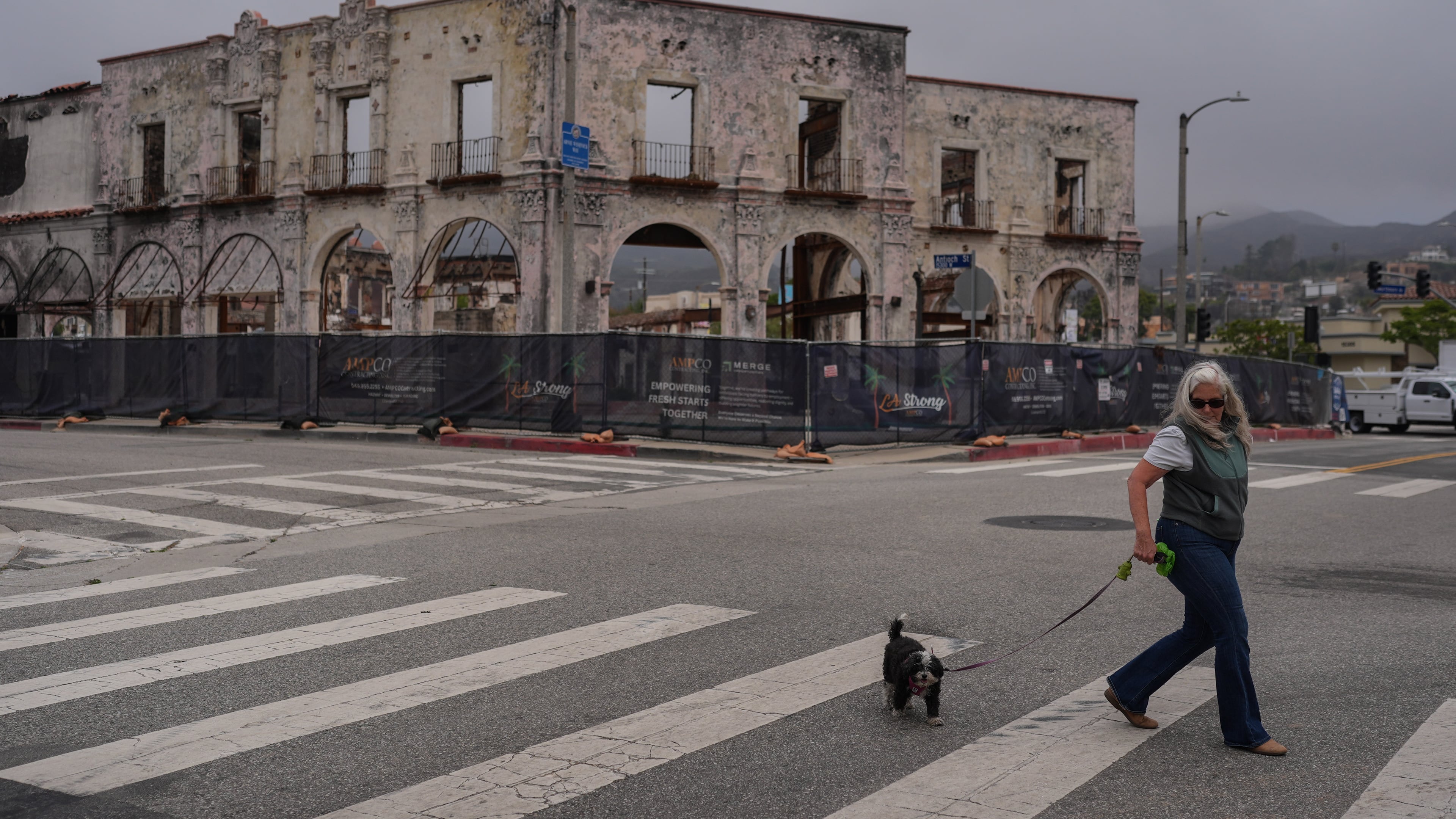 D. Berryman walks her dog, Tiny Dancer, past a fire-damaged building more than a year after the Palisades Fire in the Pacific Palisades neighborhood of Los Angeles, Thursday, June 4, 2026. (AP Photo/Jae C. Hong)