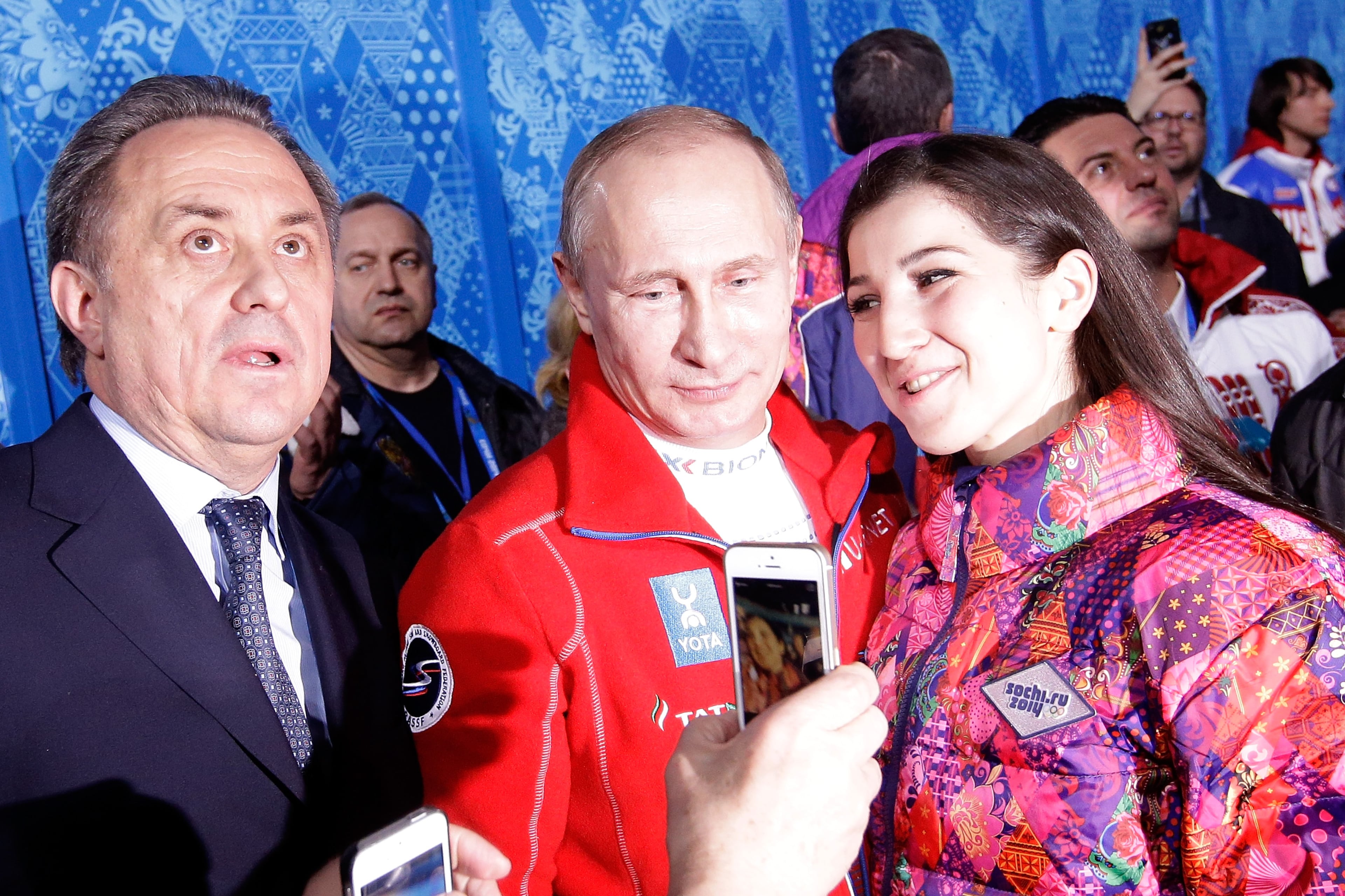 Russian Minister of Sport, Tourism and Youth policy Vitaly Mutko (L) looks on as Russian President Vladimir Putin (C) poses with a fan after the Flower Ceremony for the Team Figure Skating Overall during day two of the Sochi 2014 Winter Olympics at Iceberg Skating Palace onon February 9, 2014 in Sochi, Russia. (Photo by Darren Cummings/Pool/Getty Images)