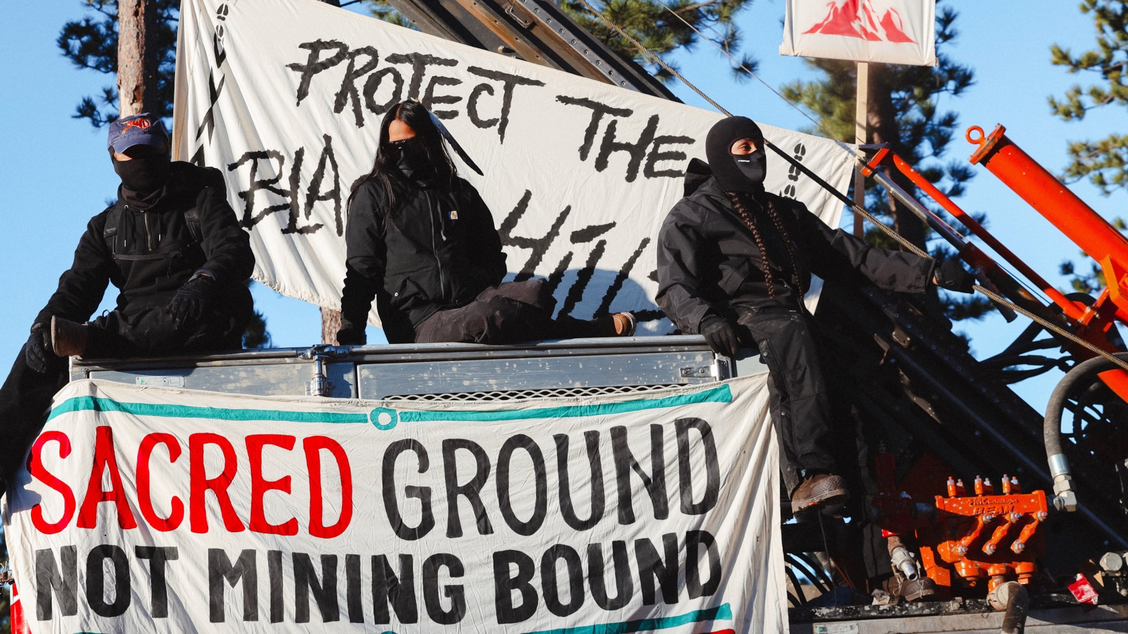 Masked demonstrators sit atop drilling equipment on Thursday, April 30, 2026, in Pennington County, S.D., with a banner reading Sacred ground not mining bound. (Angel White Eyes via AP)