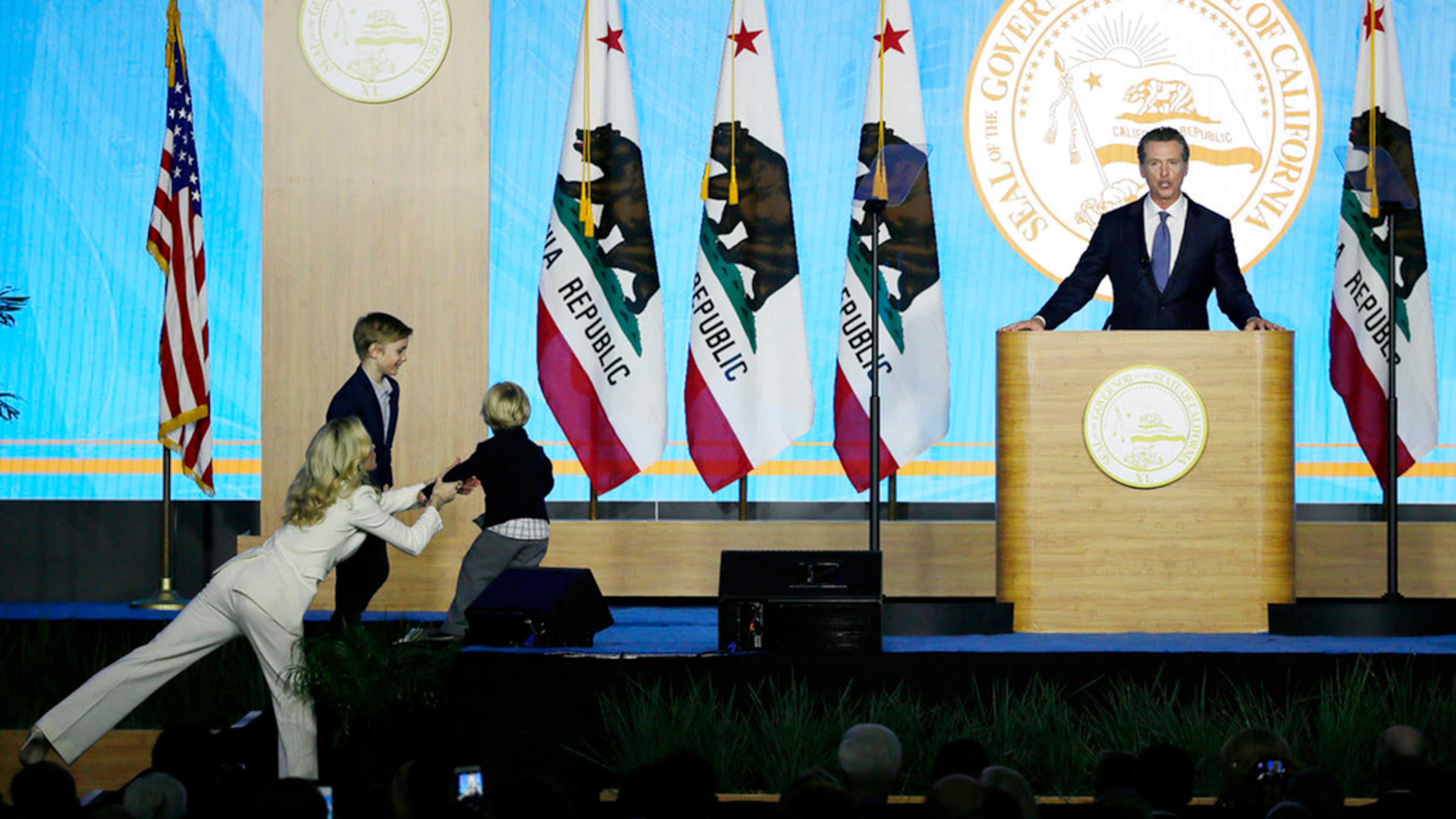 California Governor Gavin Newsom speaks during his inauguration as his wife, Jennifer Siebel Newsom, left, tries to grab their son, Dutch, who was running around the stage Monday, Jan. 7, 2019, in Sacramento, Calif. Looking on is their son Hunter.