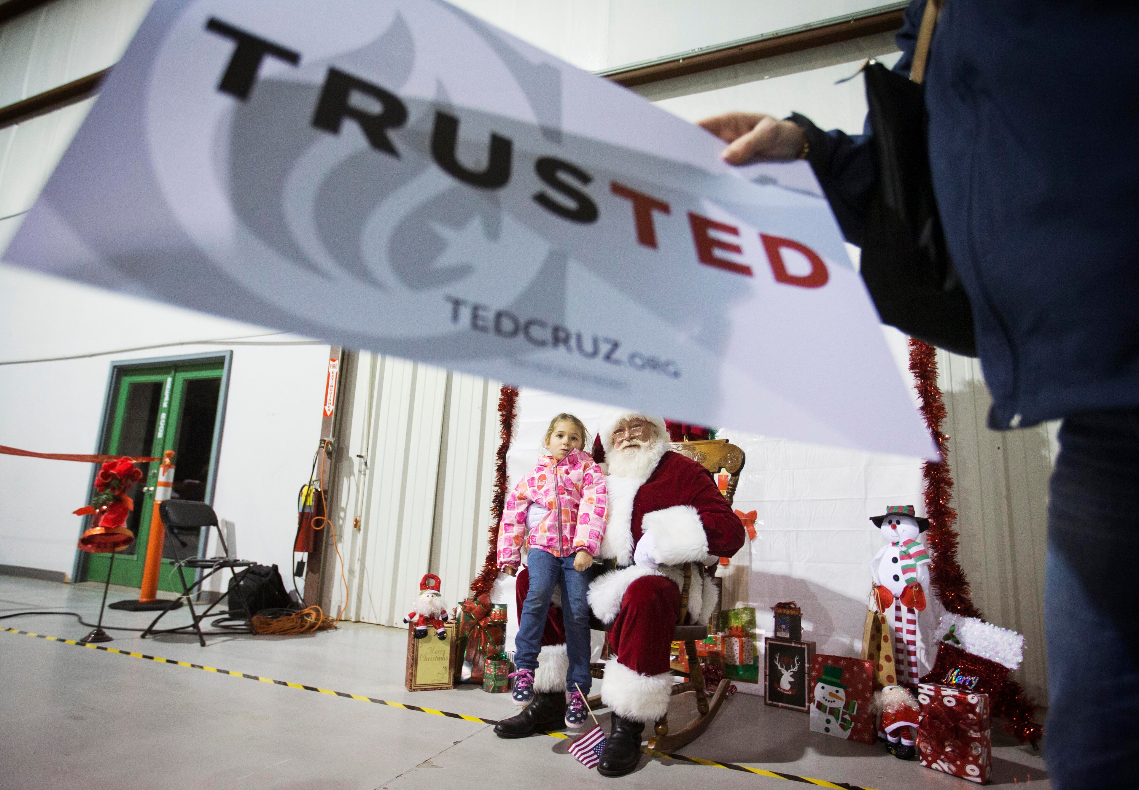 Annabelle Schaeffer, 5, left, sits on Santa Claus' lap as her mother Amy Schaeffer, right, both of Atlanta, grabs a sign while taking pictures with Santa before Republican presidential candidate, Sen. Ted Cruz, R-Texas, arrives for a campaign event Friday, Dec. 18, 2015, in Kennesaw, Ga. Cruz is on an eight-state, 12-city "Christmas Tour," and at each stop, the campaign plans to have Santa Claus on hand, posing for pictures and kissing babies. (AP Photo/David Goldman)