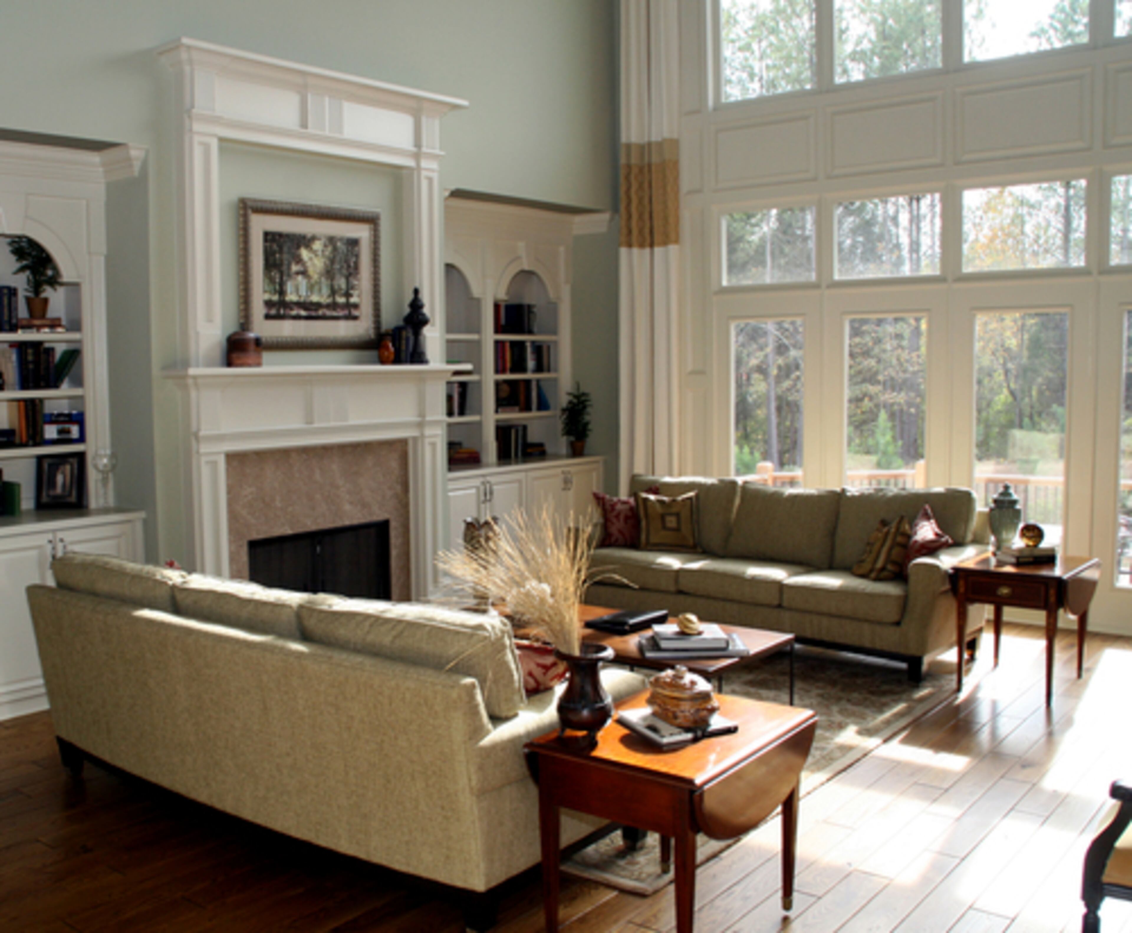 GREAT ROOM: A soaring wall of windows bathes sage-colored walls in light for an airy entertainment spot. The floor-to-ceiling window treatments are accented with a band of warm gold brocade; another layer of fabric texture comes from the linen-upholstered pair of sofas flanking the fireplace. A pair of drop-leaf end tables adds the European touch to the room.