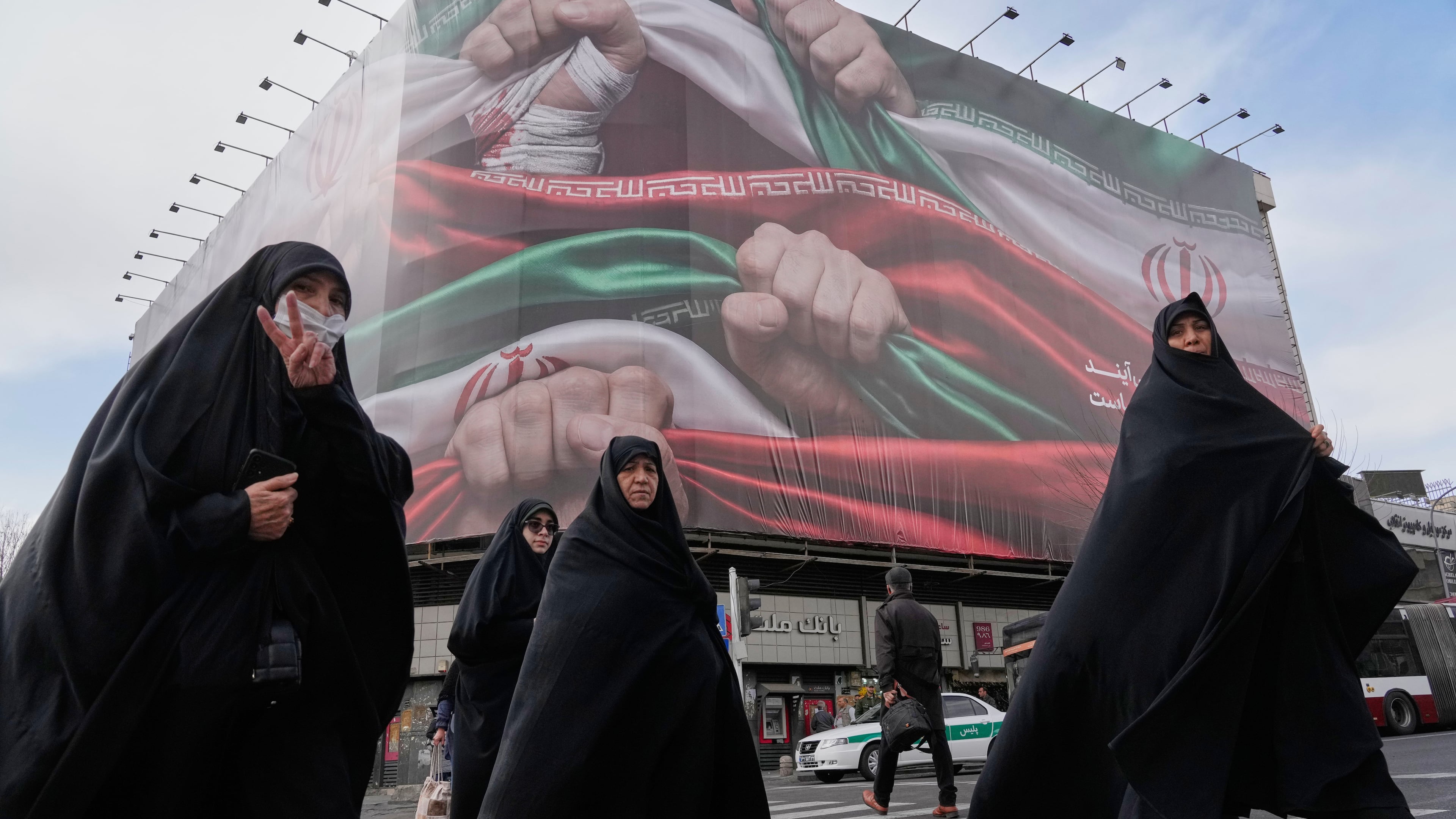 Women cross a street under a huge banner showing hands firmly holding Iranian flags as a sign of patriotism, as one of them flashes the victory sign, in Tehran, Iran, Wednesday, Jan. 14, 2026. (AP Photo/Vahid Salemi)