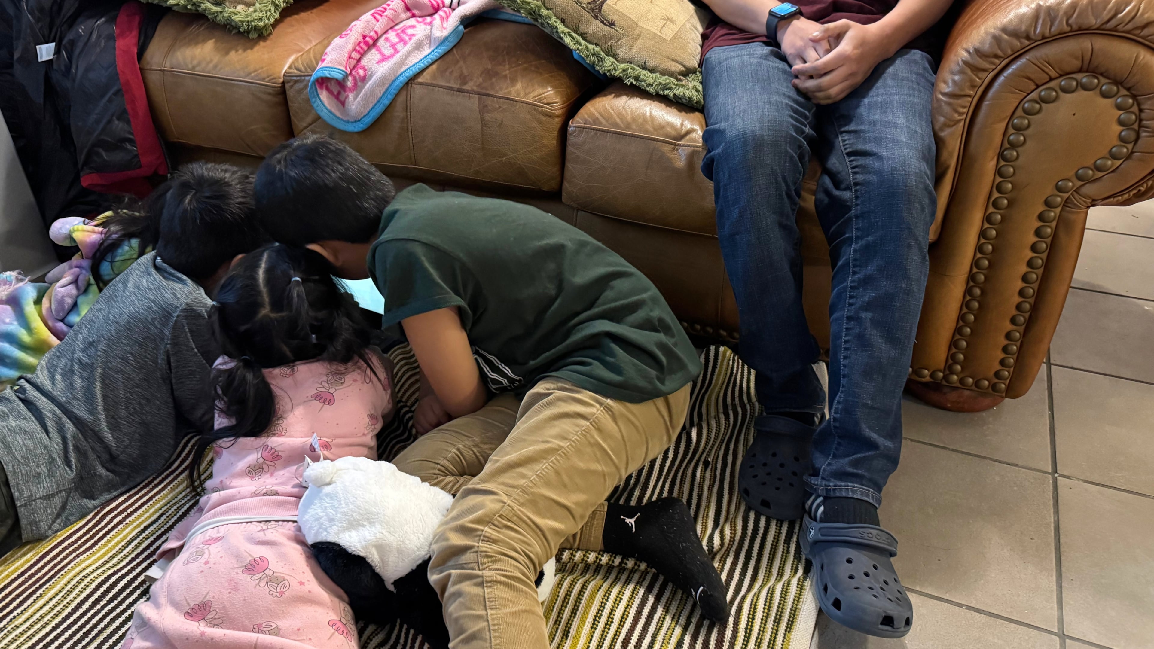 A boy sits on the couch of a Minneapolis safe house Friday, Jan. 16, 2026, as his younger siblings and niece play on the floor beside him after they fled their home because they were sought by federal immigration enforcement agents. (AP Photo/Jack Brook)