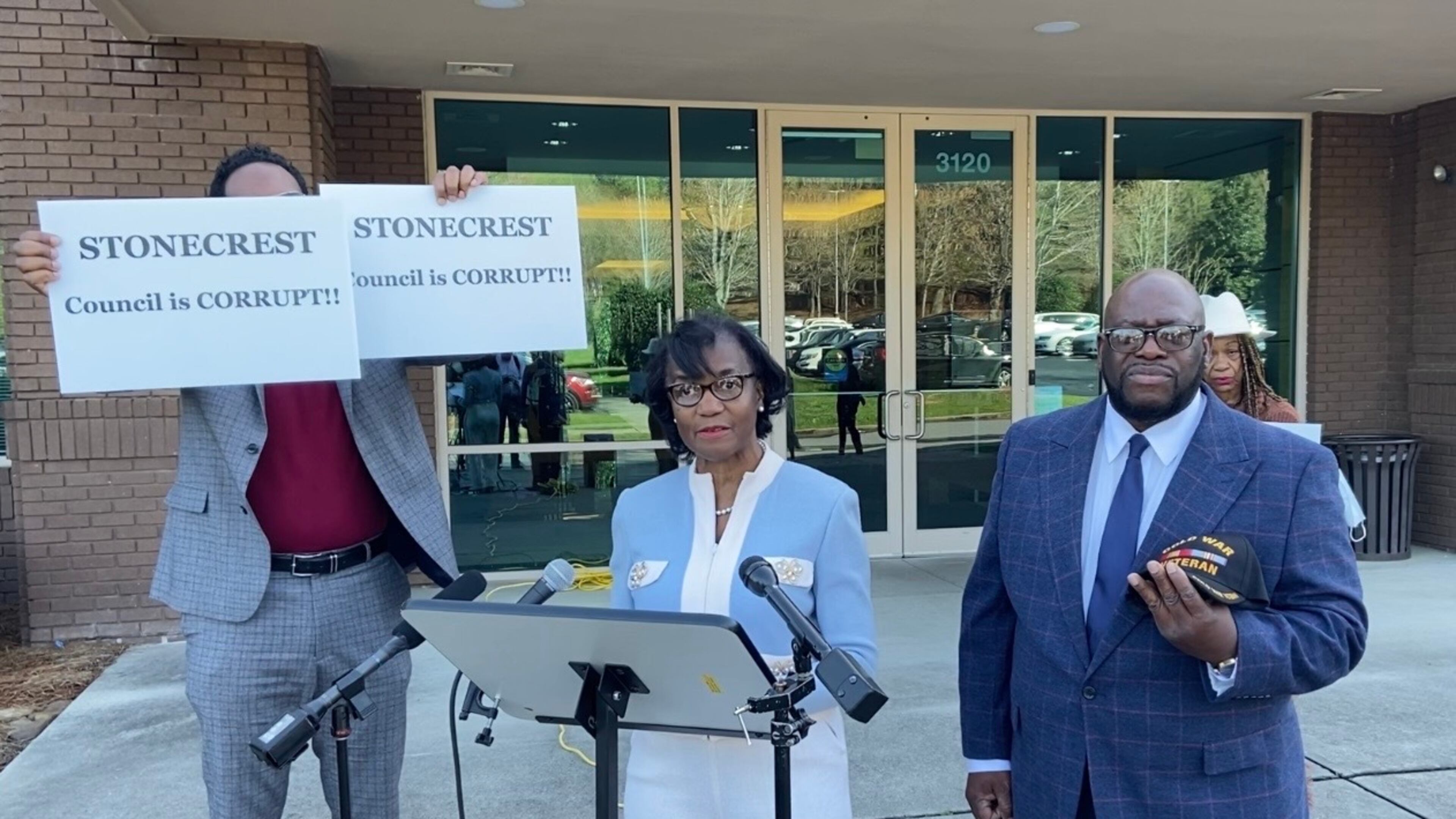 Michael Fayoyin, left, holds signs that read, "Stonecrest Council is Corrupt!!" during the news conference held by mayoral candidates Diane Adoma (center) and Kirby Frazier (right).