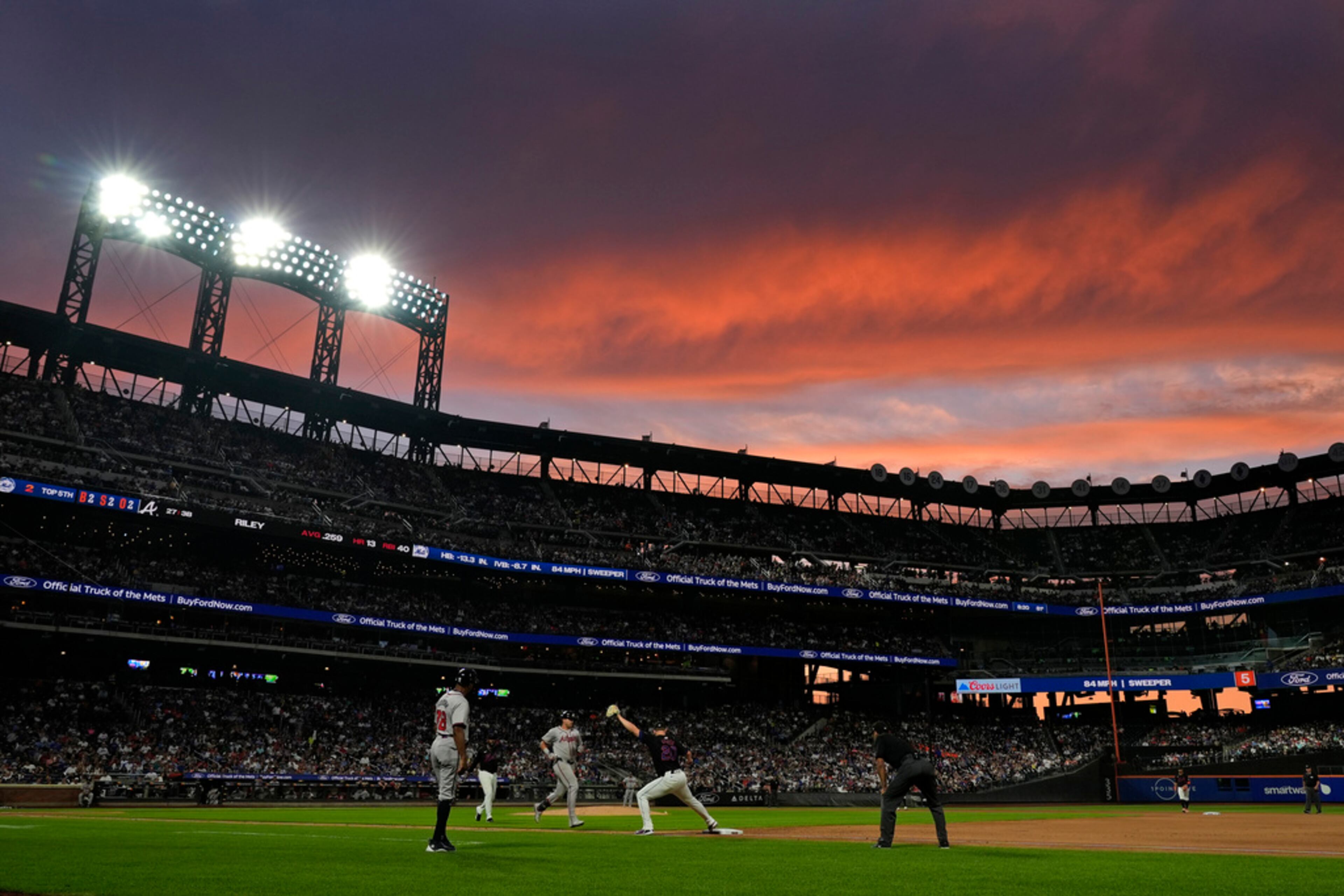 The setting sun illuminates the sky as Atlanta Braves' Austin Riley, center, is forced out at first base as New York Mets first baseman Pete Alonso catches the ball thrown by catcher Francisco Alvarez during the fifth inning of a baseball game, Thursday, July 25, 2024, in New York. (AP Photo/Pamela Smith)